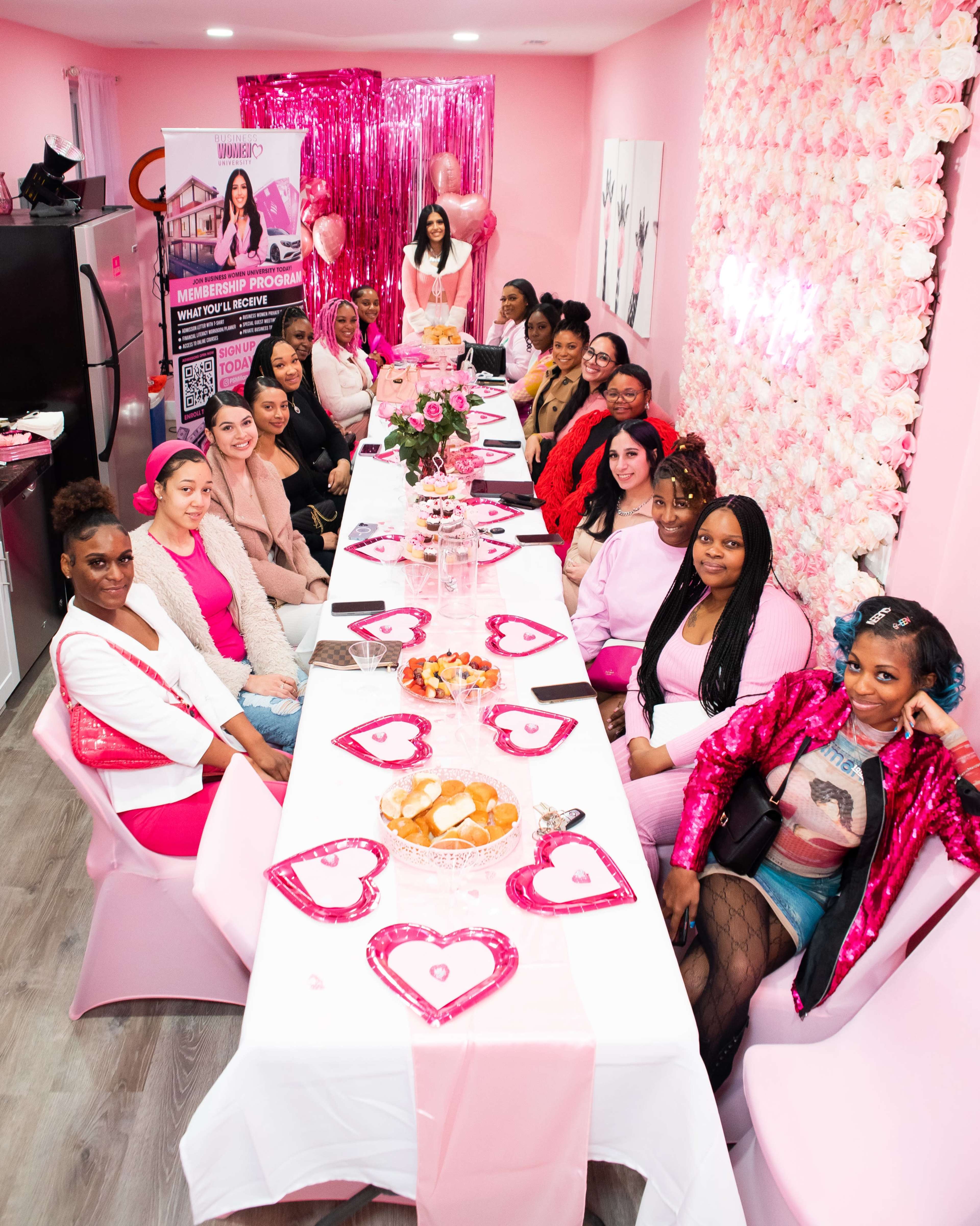 A group of women sit around a long table adorned with pink decorations and heart-shaped plates in a brightly colored room.