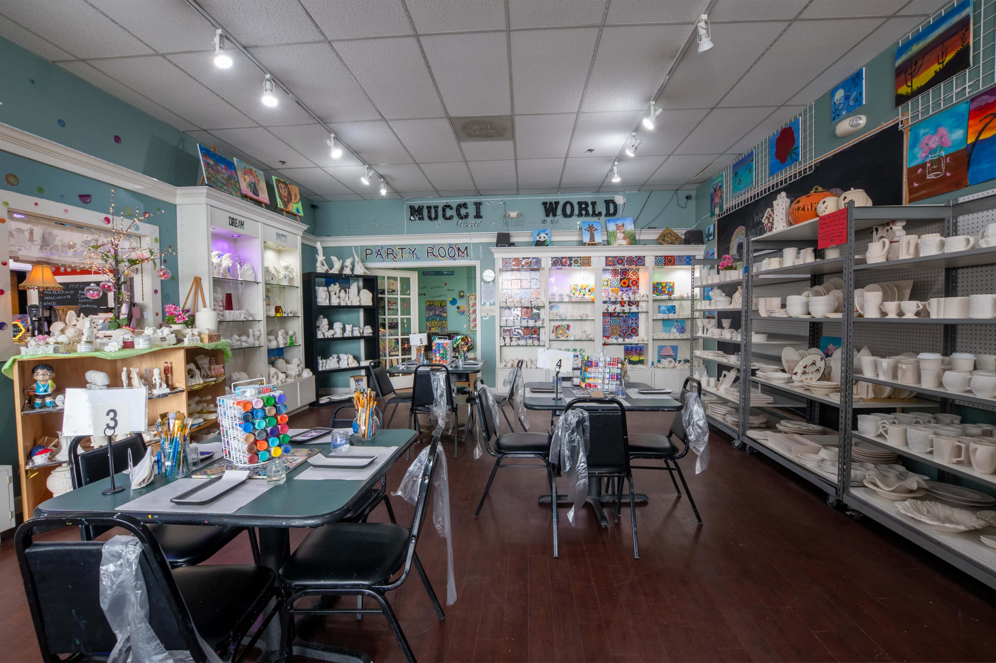 A brightly lit arts and crafts studio with tables set for painting, surrounded by display shelves filled with ceramic items and various art supplies.