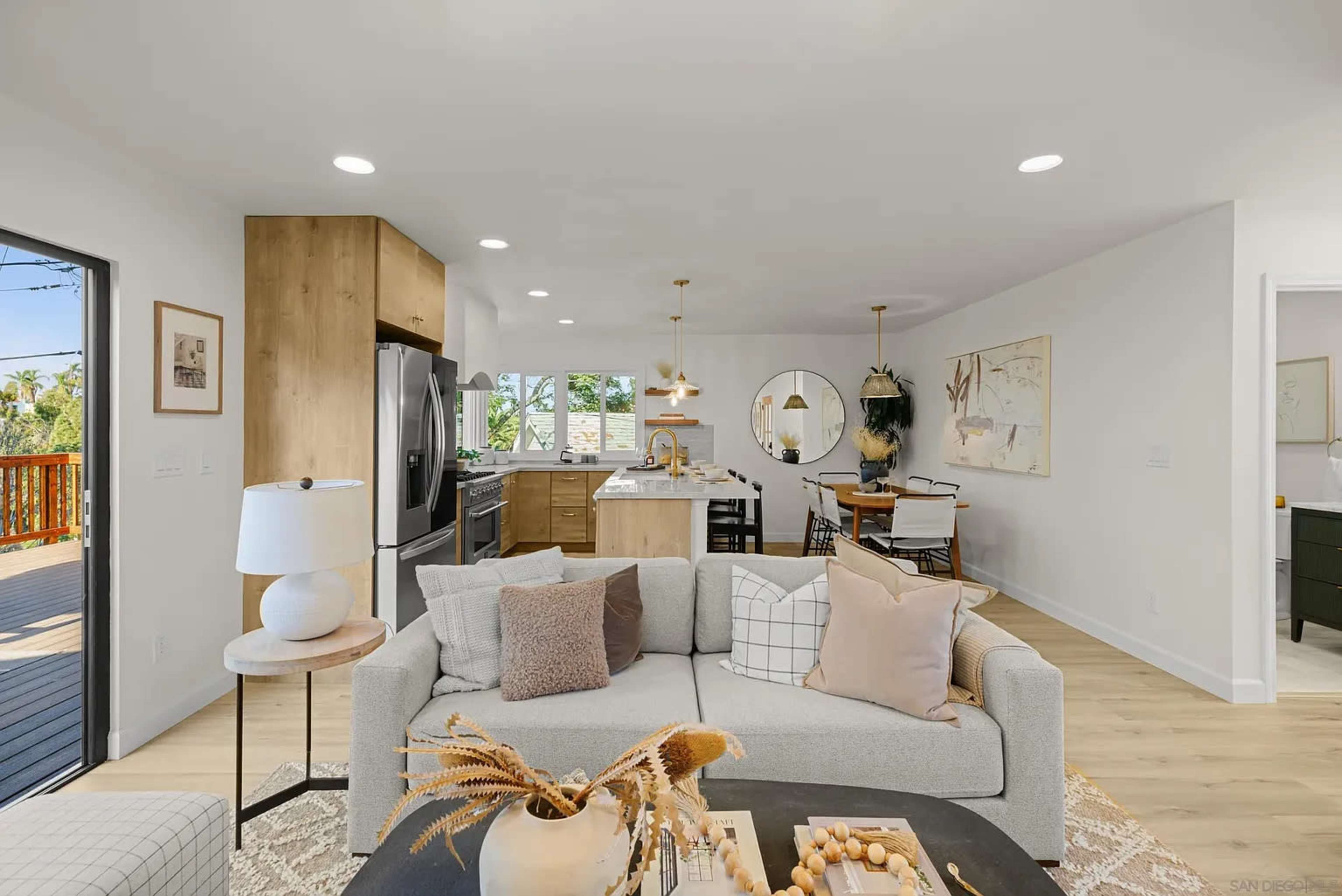 The image shows a modern, open-concept living area with a sofa, kitchen, and dining space featuring light-colored furnishings and wood accents.