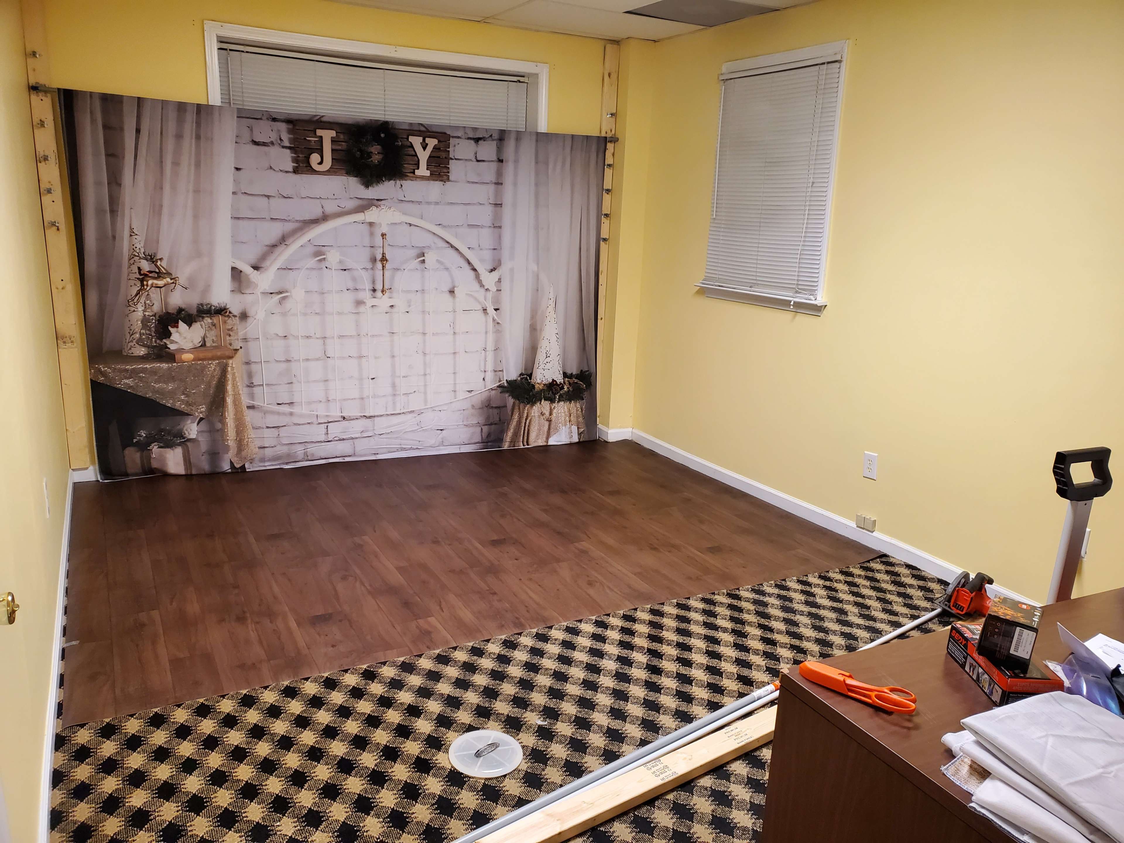 An empty room with a yellow wall, featuring a backdrop with a white arch and decorations, set up on a wooden floor next to a patterned carpet and some tools on a desk.