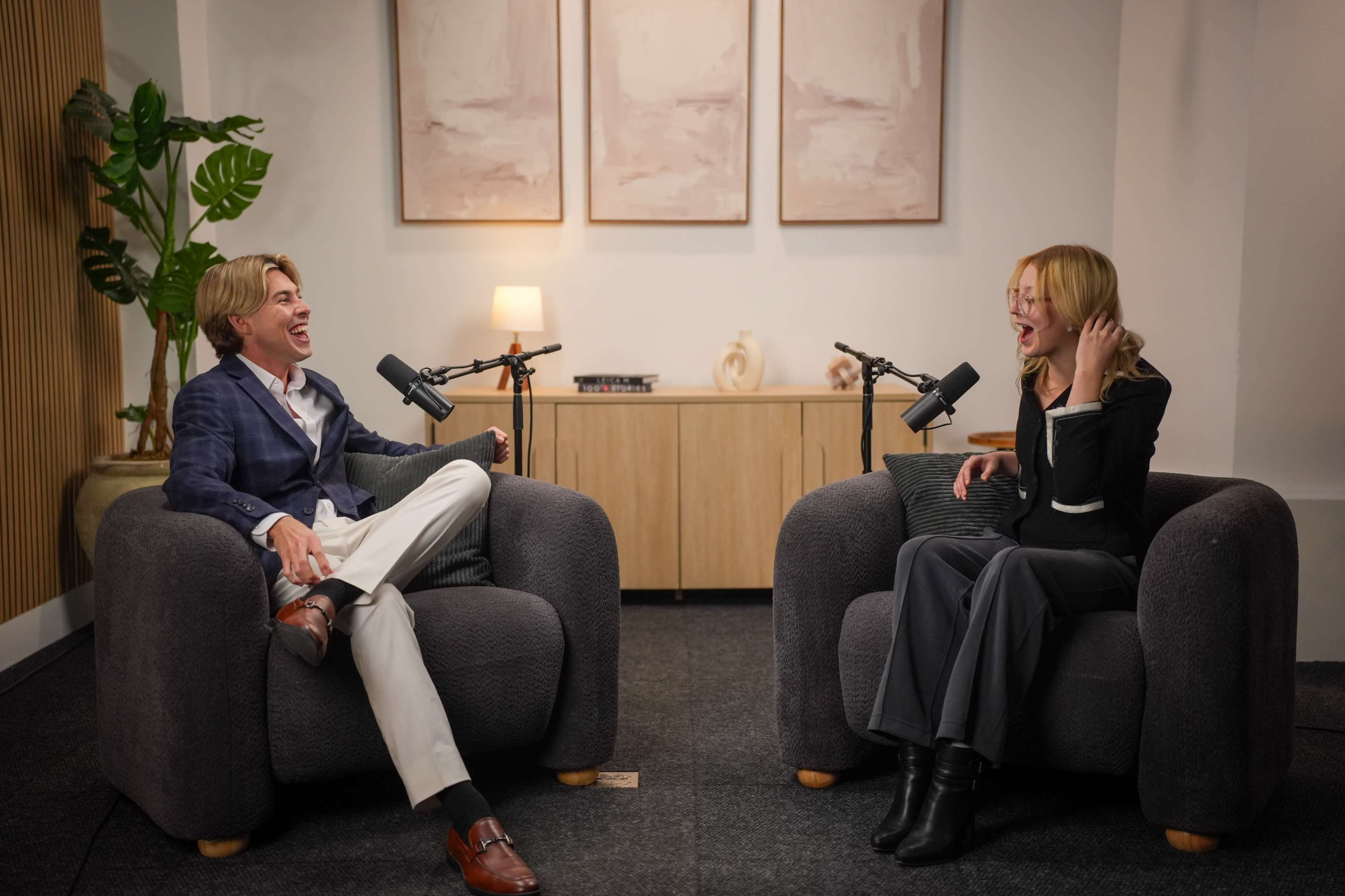 Two individuals are seated in cozy armchairs, engaging in conversation with microphones positioned between them in a well-lit, modern setting.