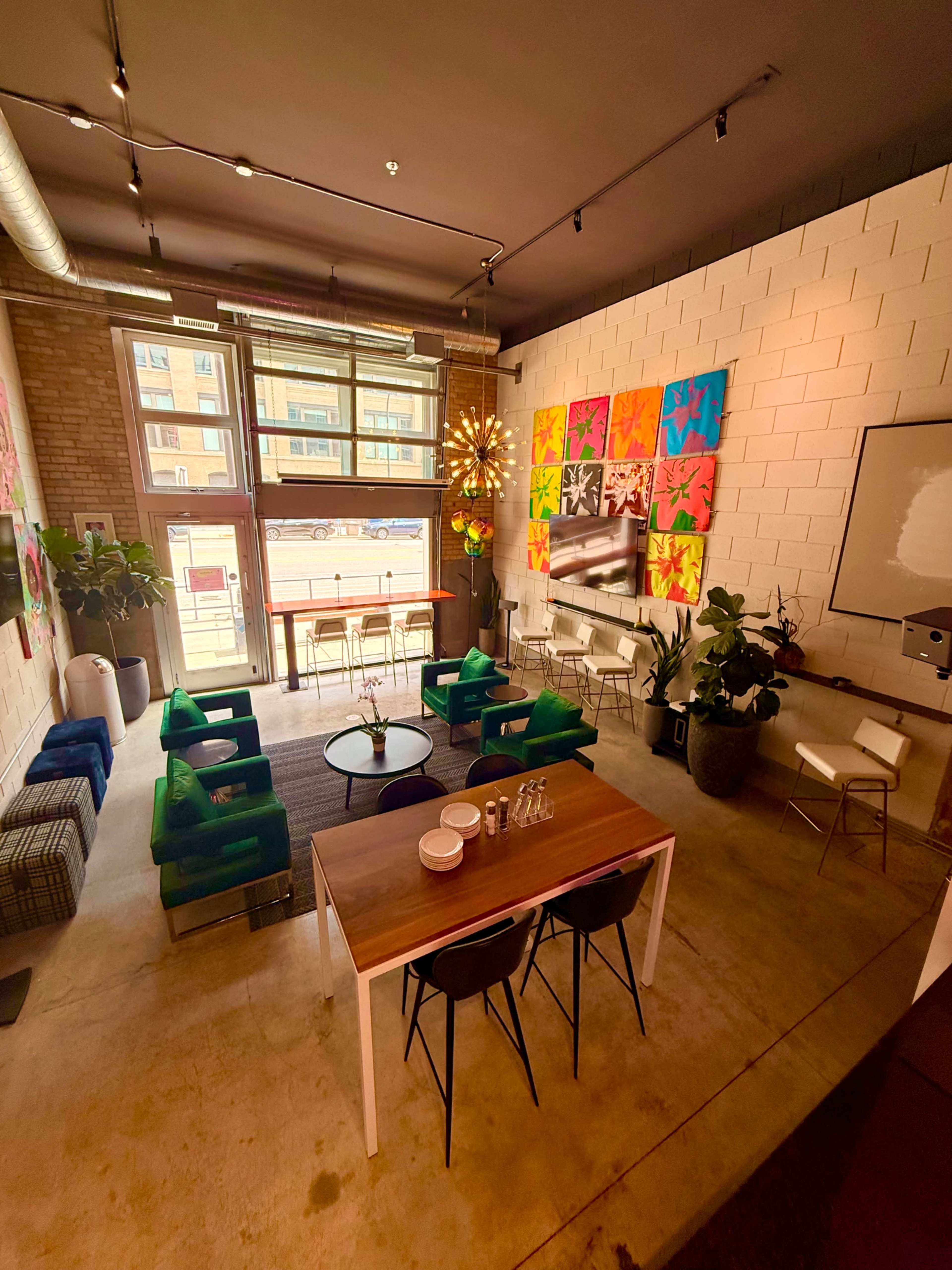 The image shows a modern indoor space with a dining table, colorful artwork on the walls, seating areas, and large windows allowing natural light to enter.