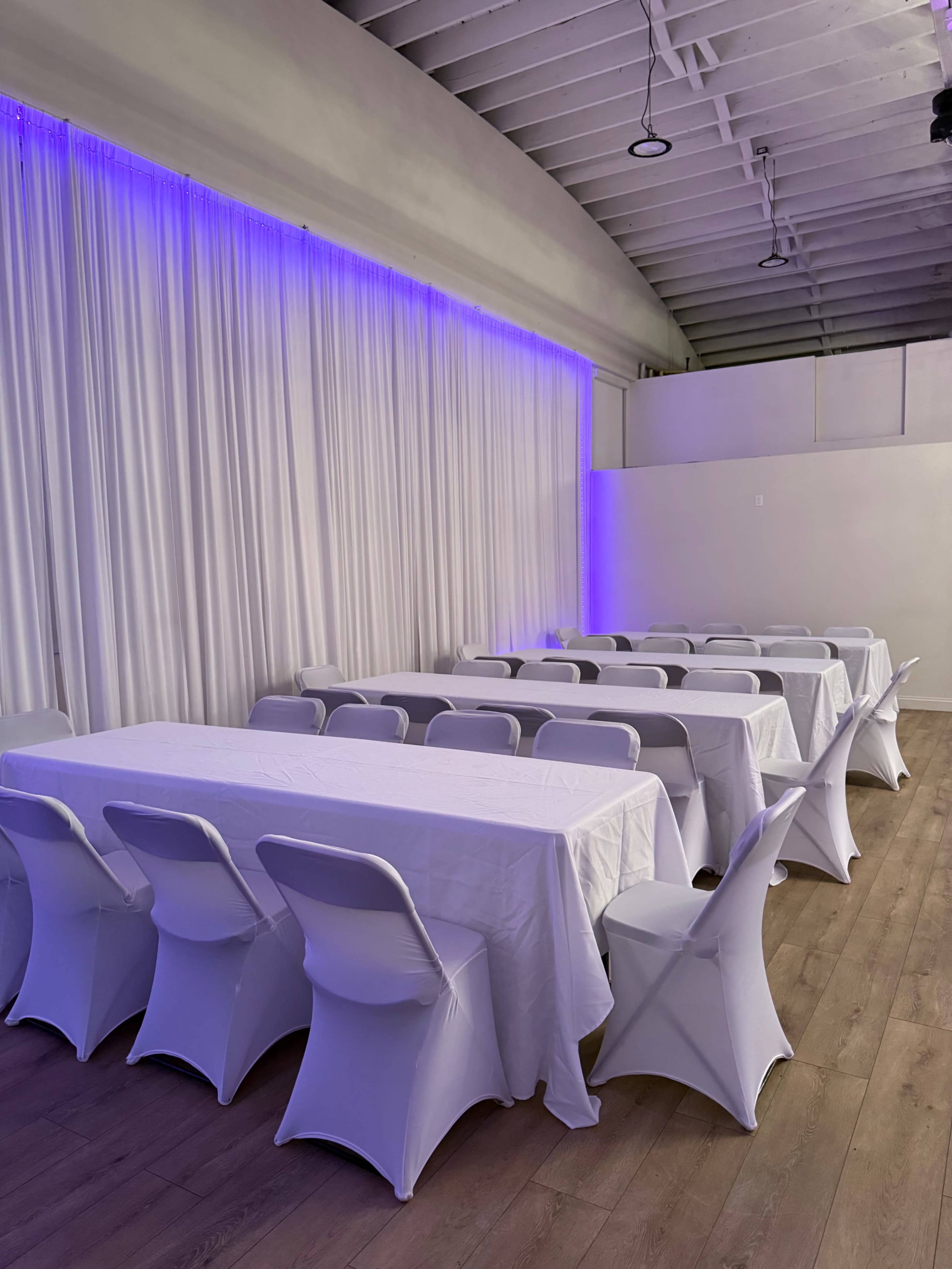 The image shows a room with multiple long tables covered in white tablecloths and white chairs, set up under soft purple lighting against a backdrop of draped white fabric.