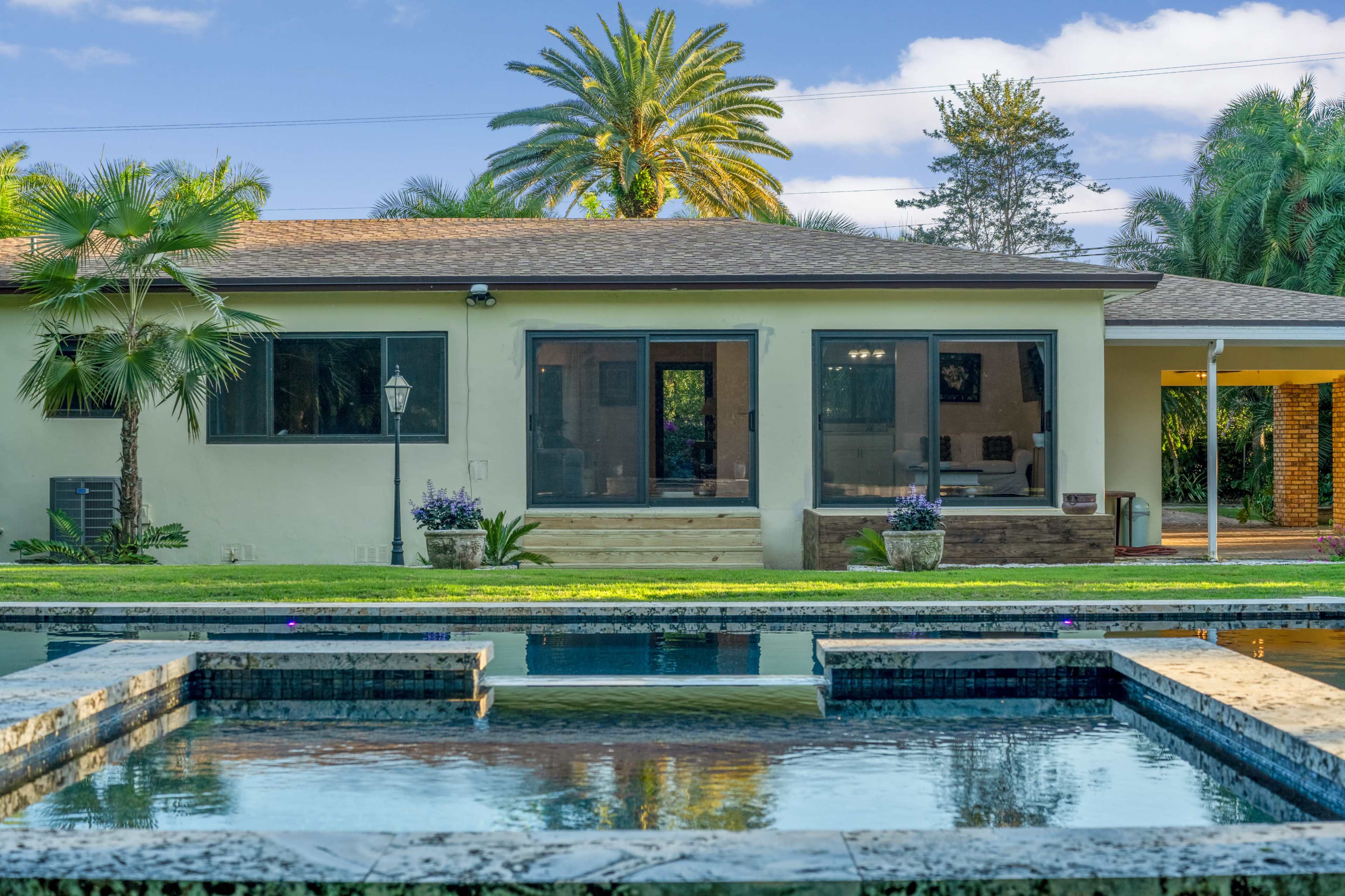 A backyard featuring a swimming pool with a spa area, surrounded by lush greenery and a single-story home in the background.