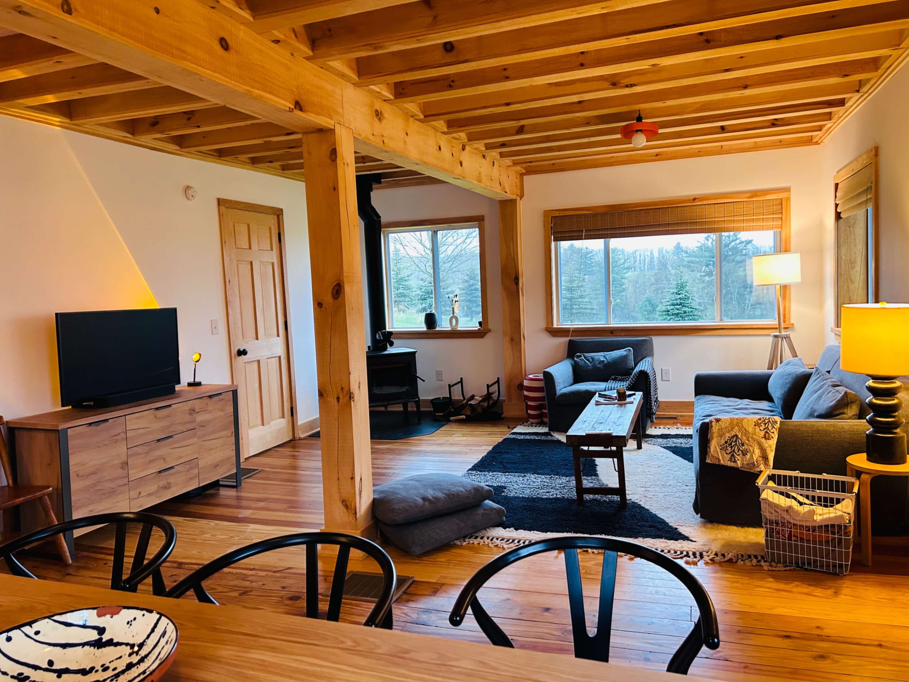 A cozy living room with wooden beams, a television, a coffee table, and a large window overlooking a wooded area.