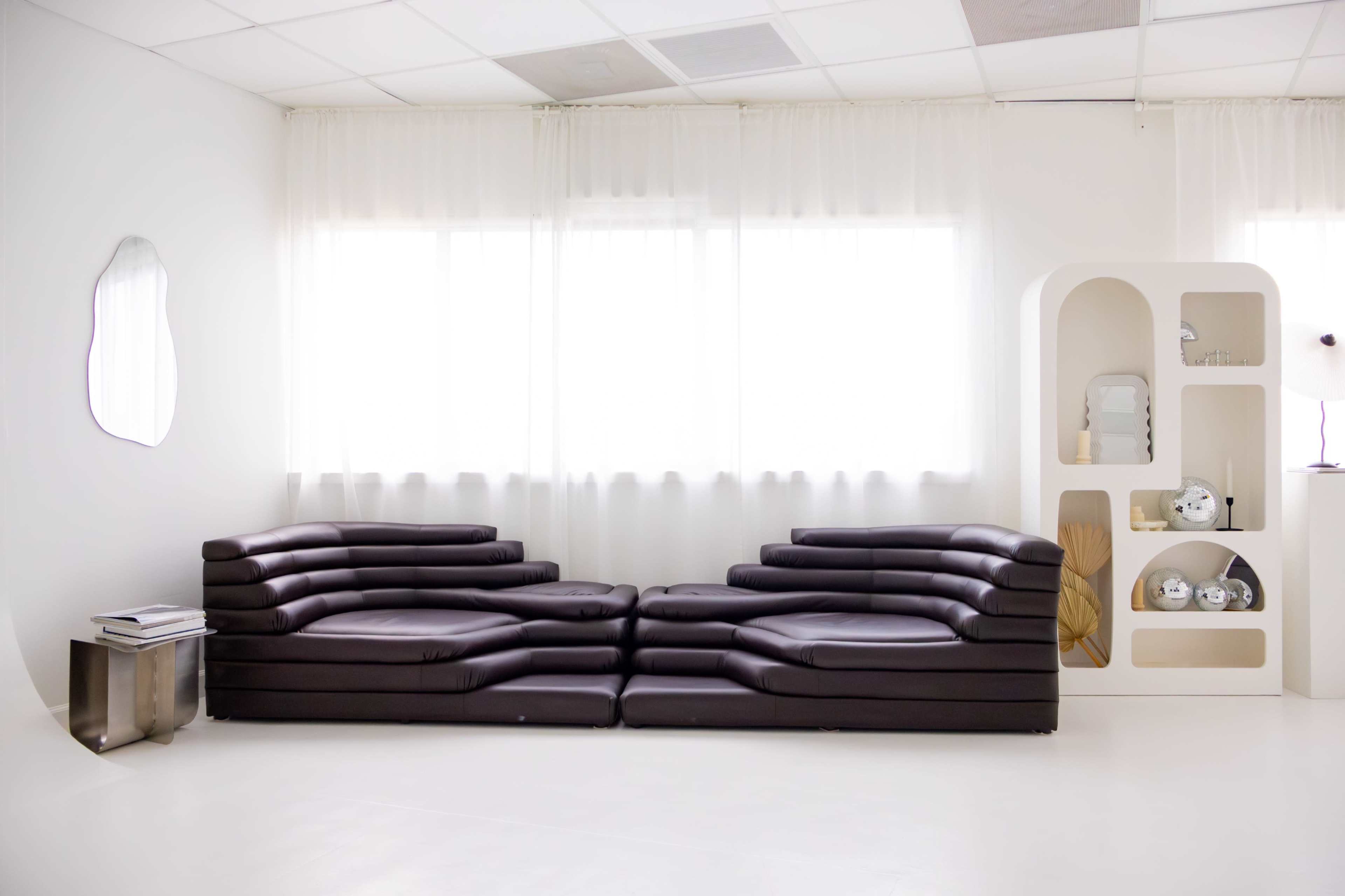 A modern, minimalist living space features a dark leather sectional sofa and a white shelving unit with decorative items against a bright, airy backdrop of large windows.