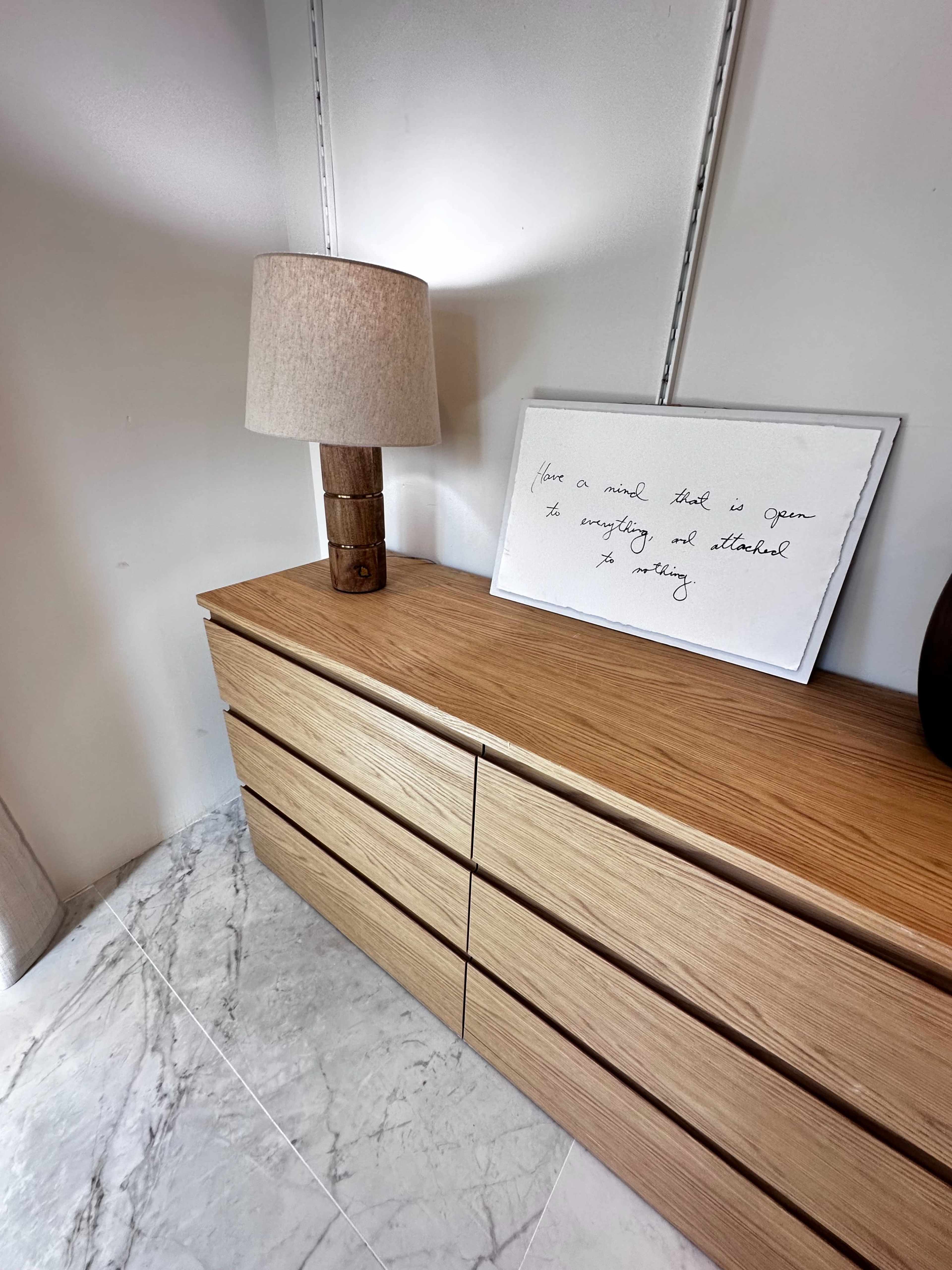 A wooden dresser with multiple drawers sits against a wall, accompanied by a textured lamp and a framed piece of art with handwritten text.