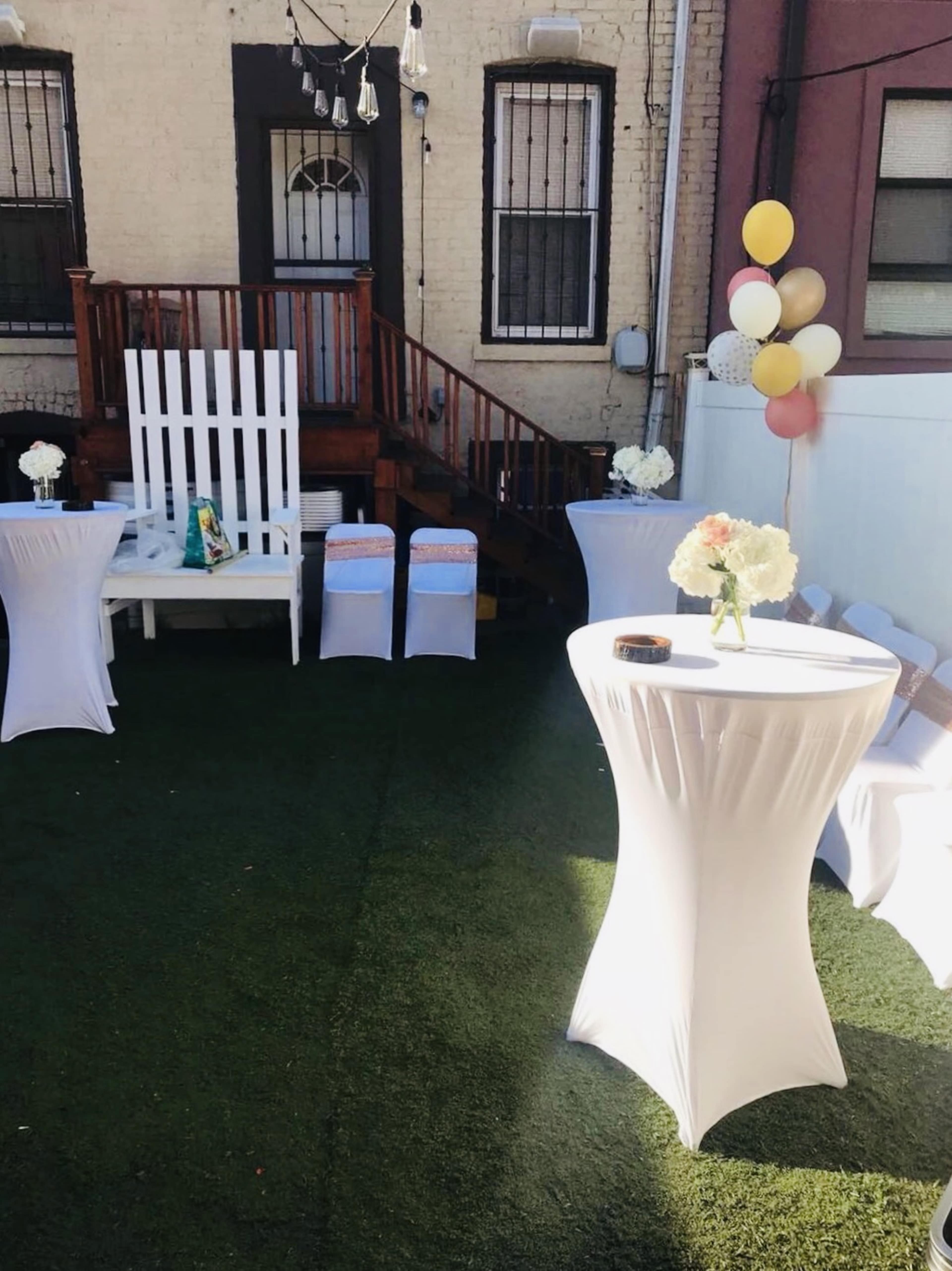 A decorated outdoor space featuring white tables, chairs, a wooden staircase, and a backdrop of balloons.