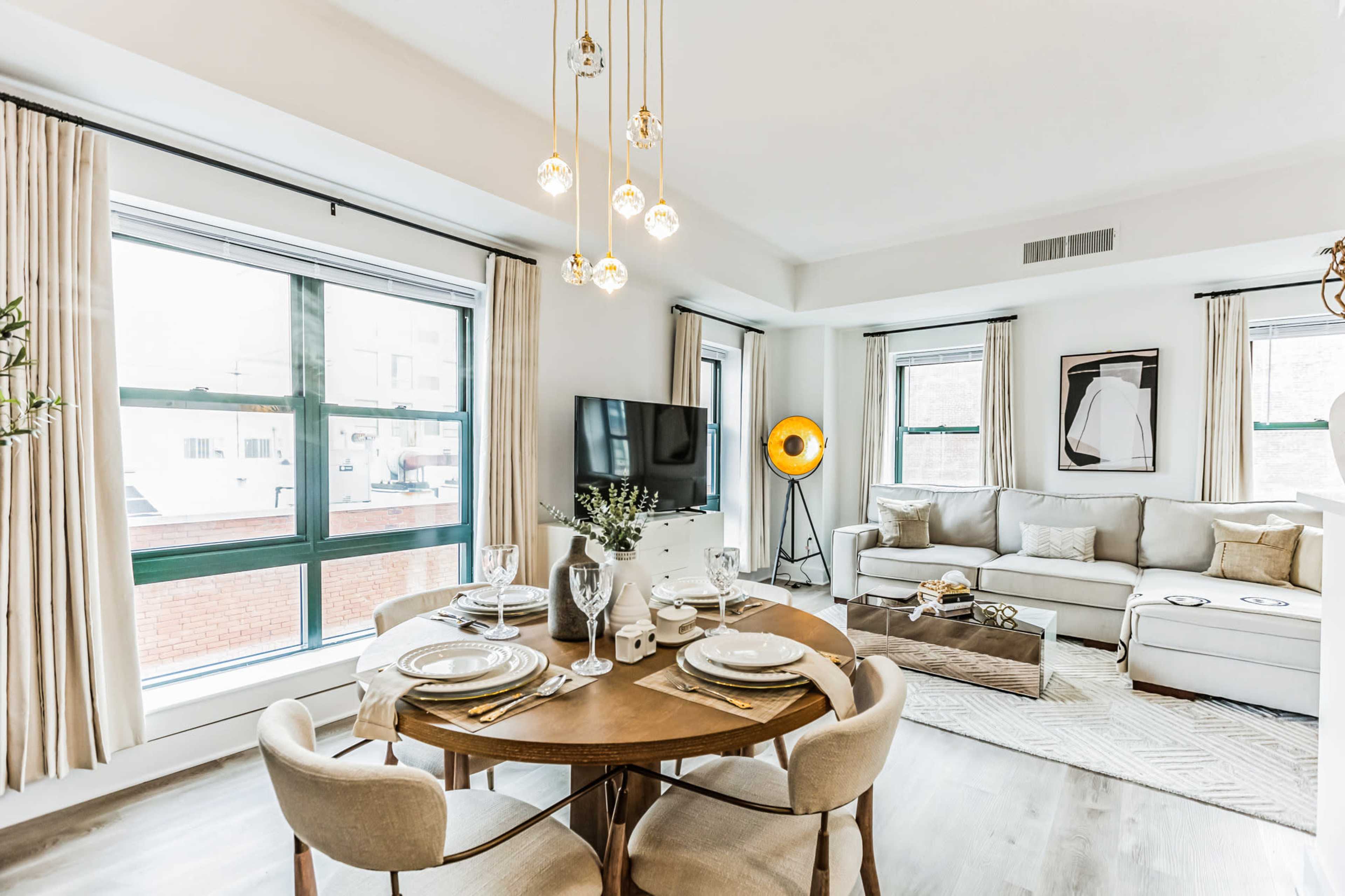 A modern living room and dining area feature a circular wooden table set for a meal, with a sofa and television in the background.