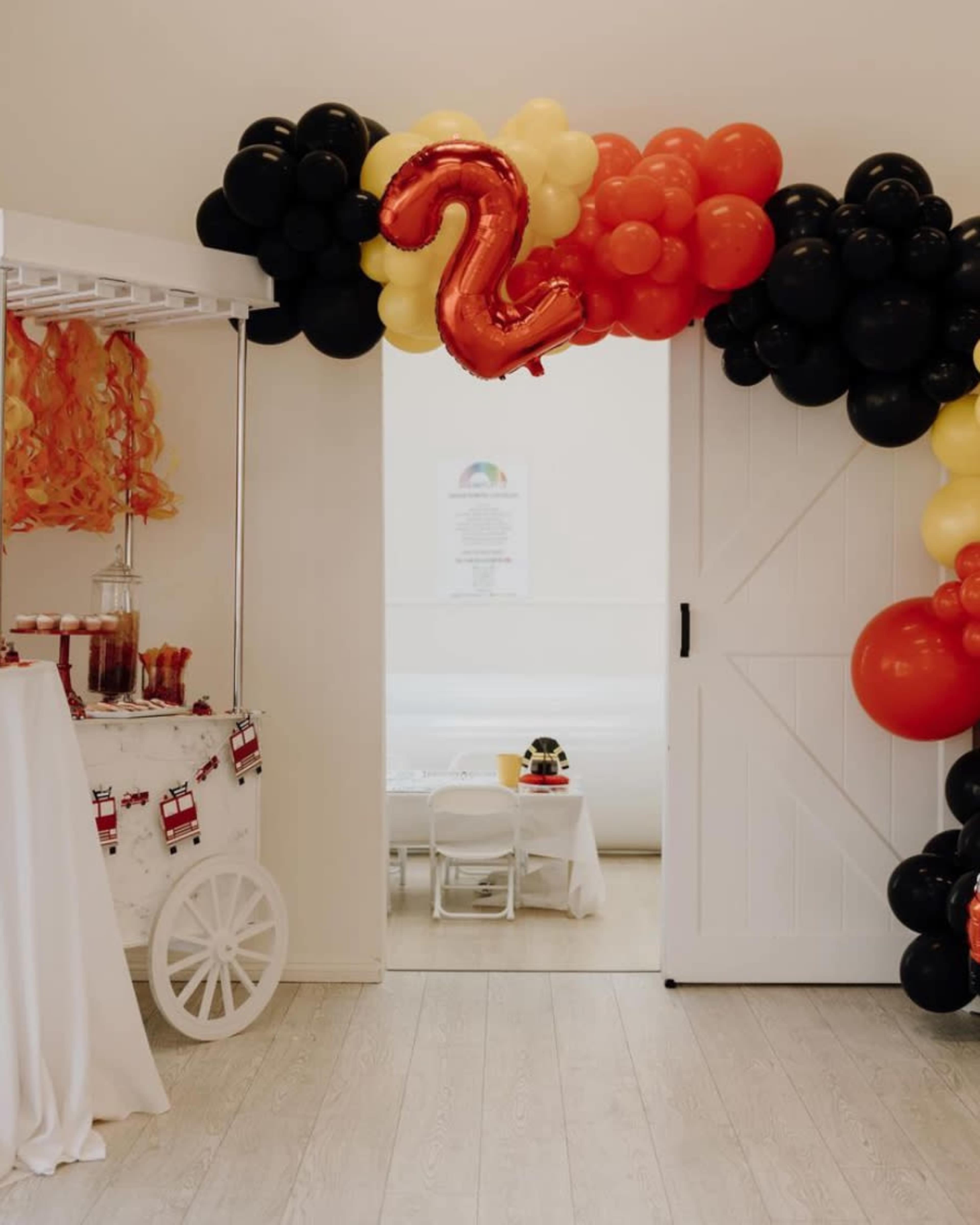 The scene features a birthday party entrance decorated with a balloon arch in black, red, and yellow, and a large red "2" balloon.