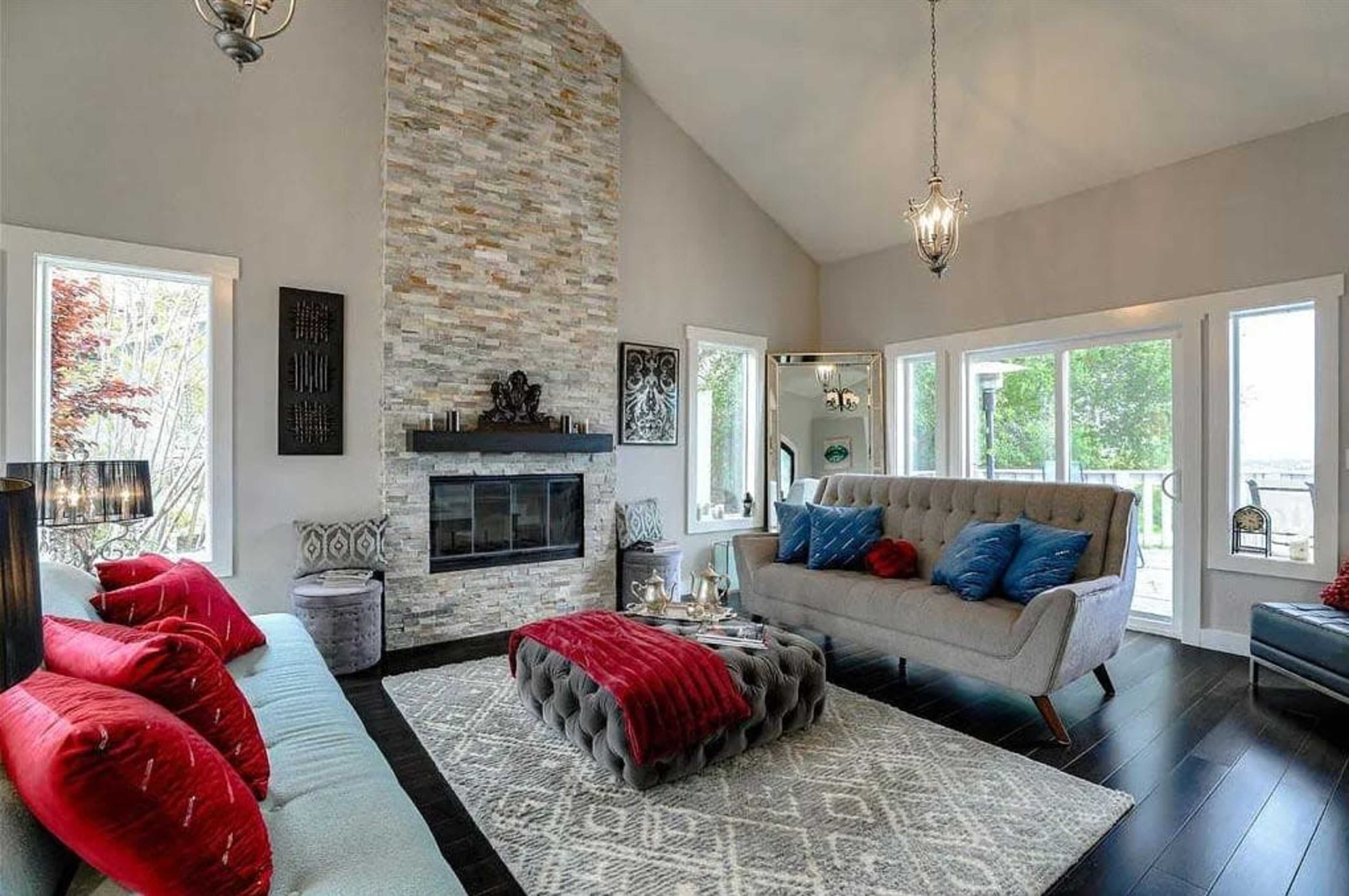 The image depicts a modern living room featuring a high stone fireplace, a light gray sofa, and large windows overlooking a green area.