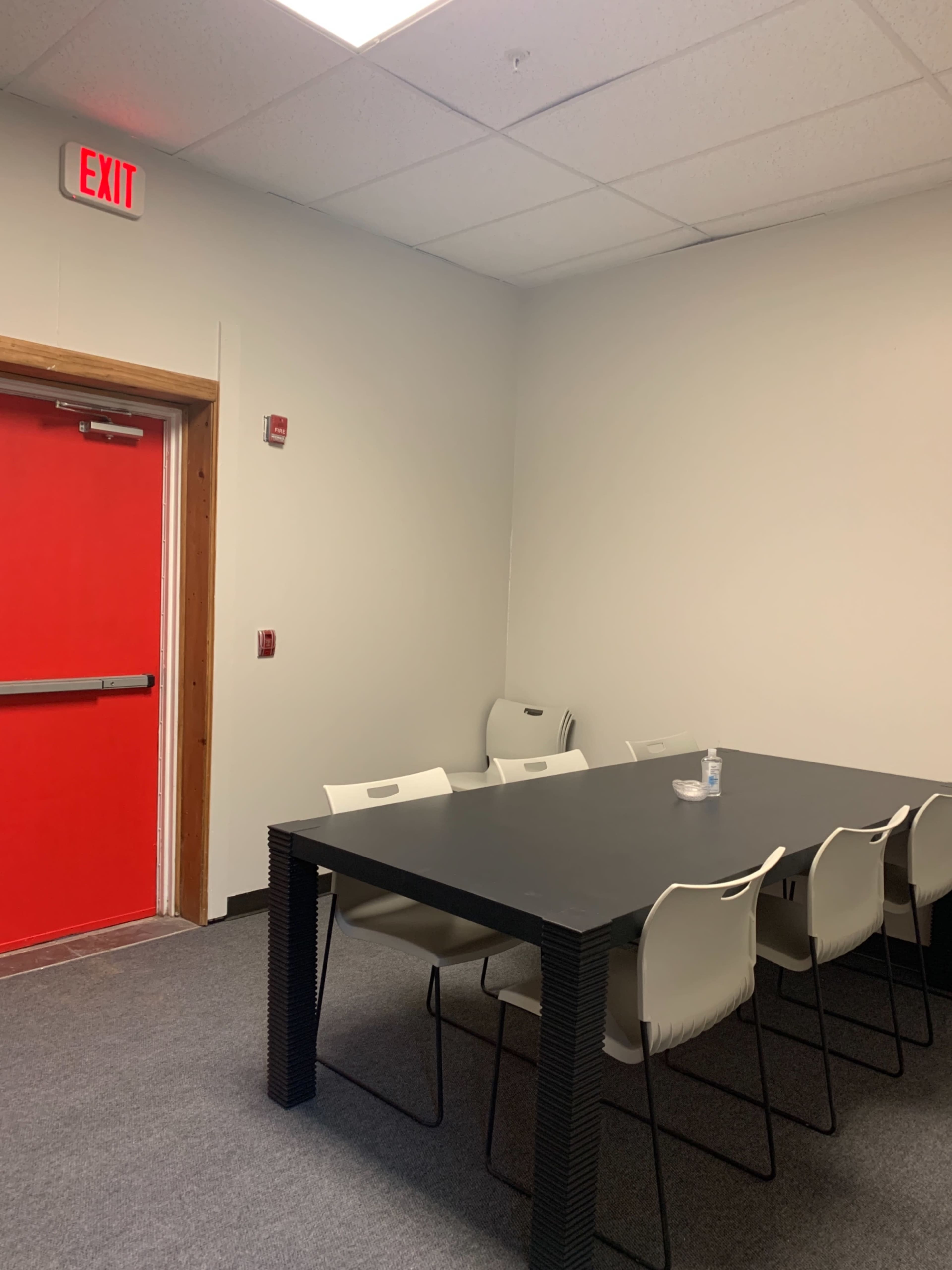 A small meeting room featuring a large black table surrounded by white chairs, with a red exit door on one wall.