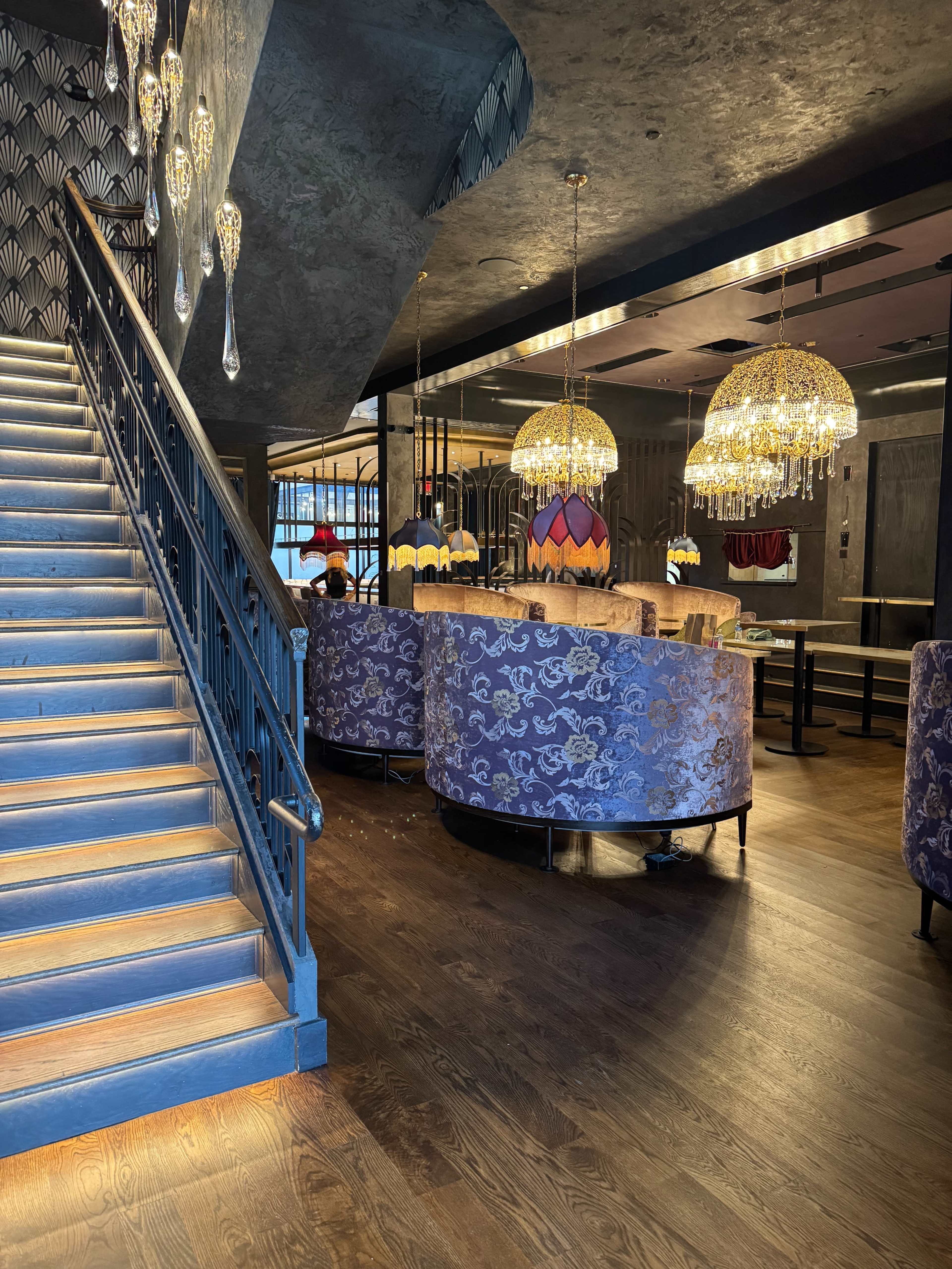 The image shows a stylish restaurant interior with plush purple seating, ornate chandeliers, and a staircase leading to an upper level.