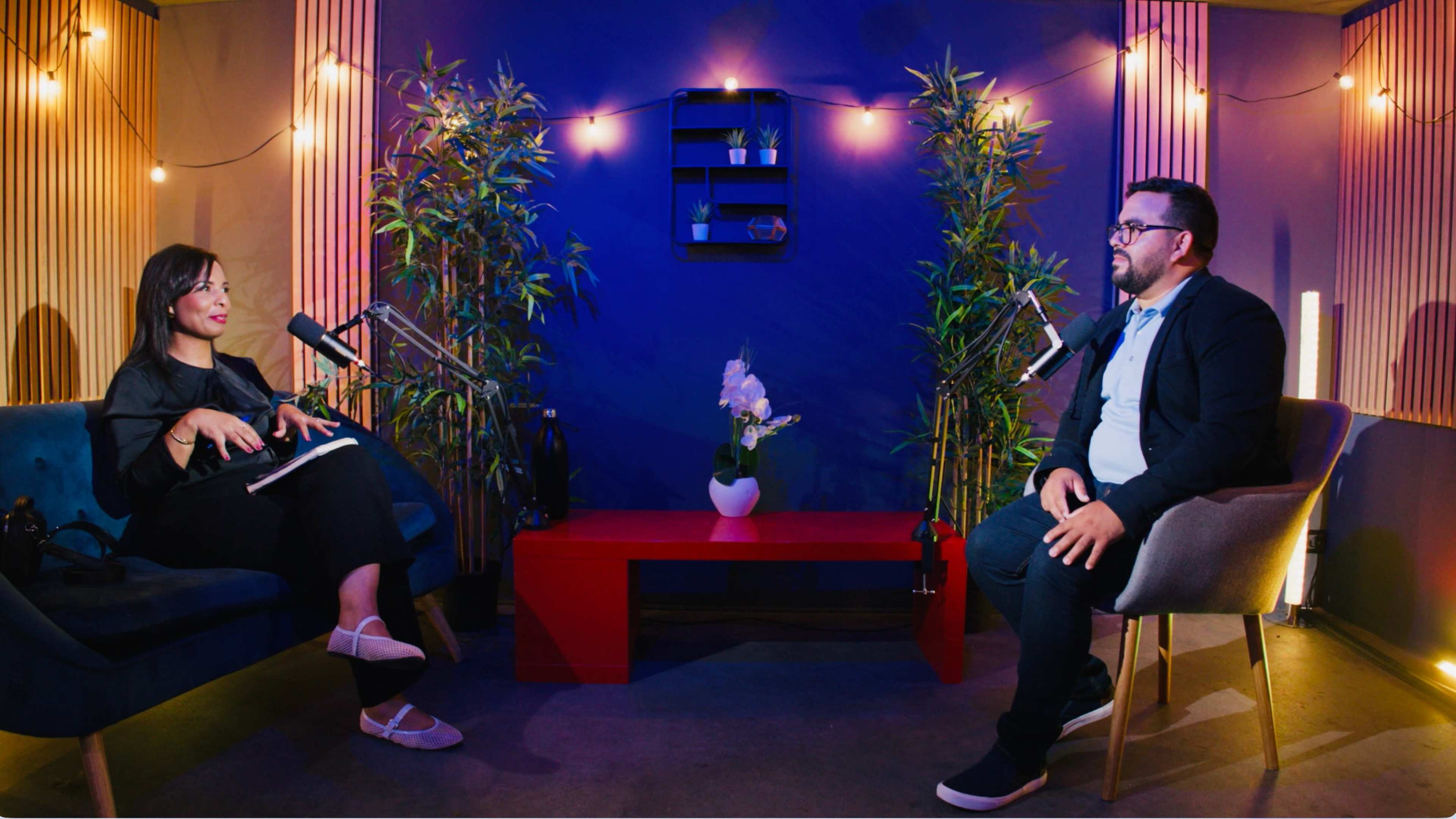 A woman and a man sit in a modern studio setting with plants and decorative lighting, engaged in conversation while microphones are positioned nearby.