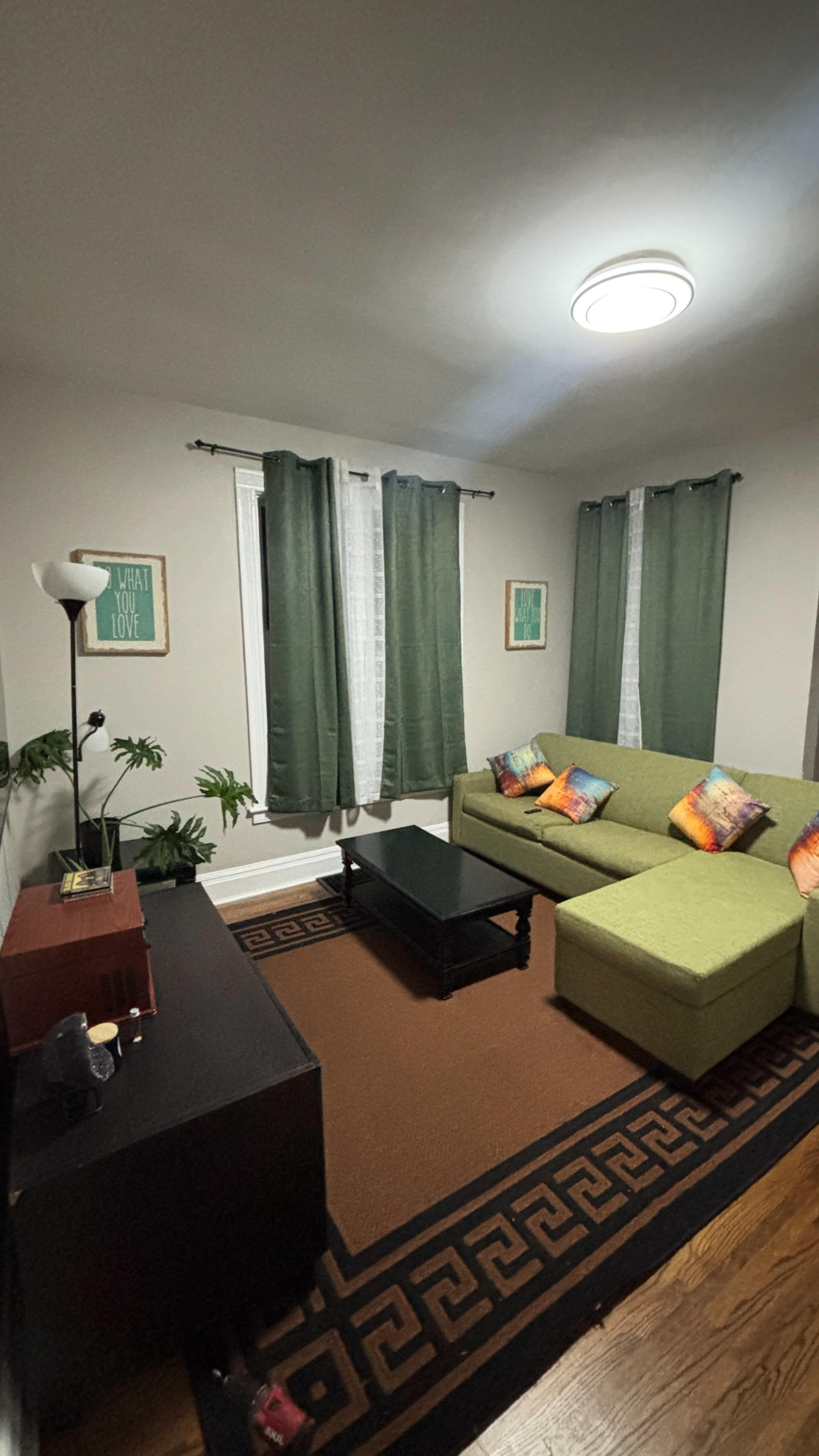 The image shows a cozy living room with a green sectional sofa, a dark coffee table, and a plant beside a window adorned with green curtains.
