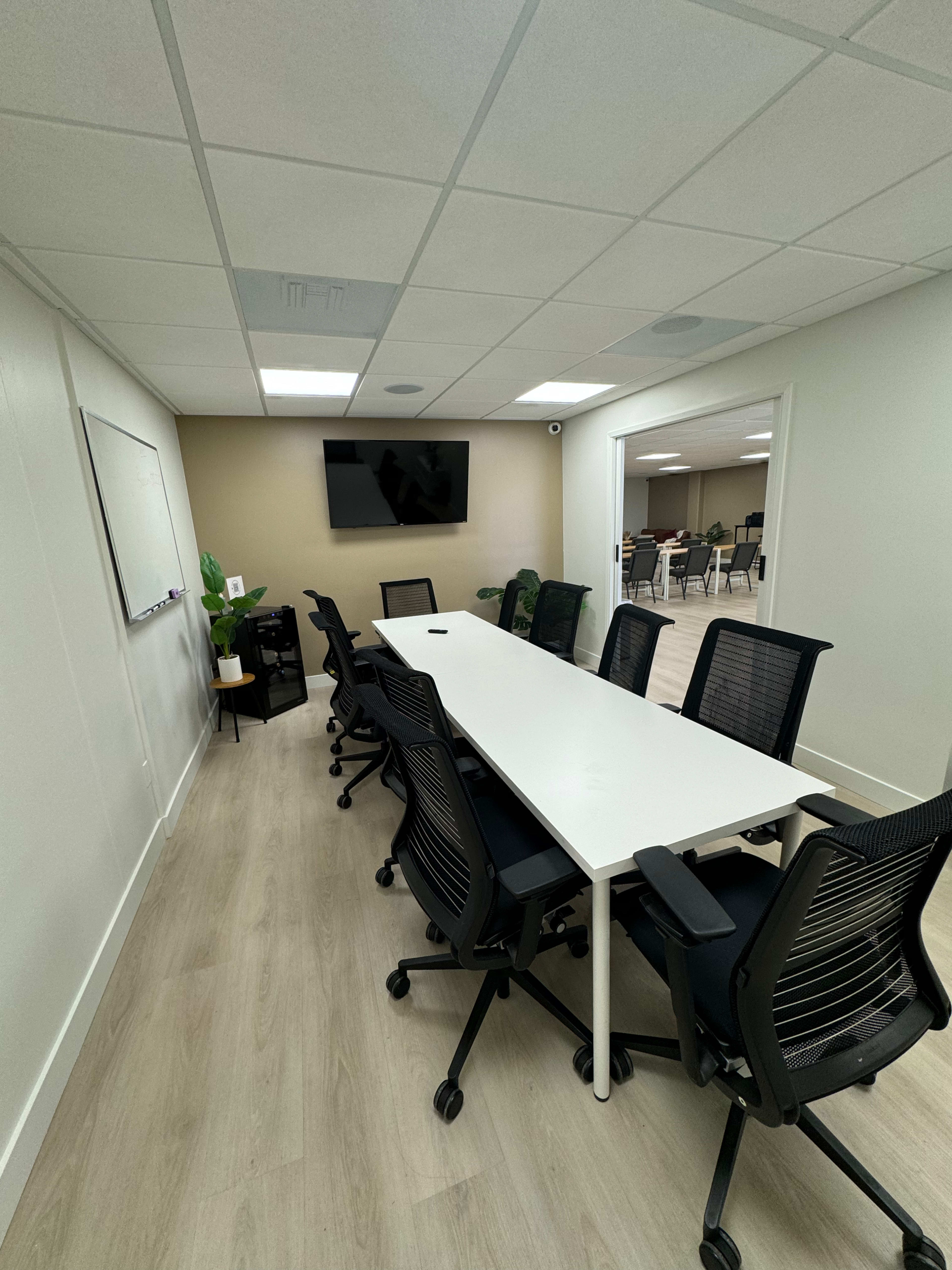 A clean, modern conference room features a long white table surrounded by black ergonomic chairs, with a television mounted on one wall and a whiteboard on another.