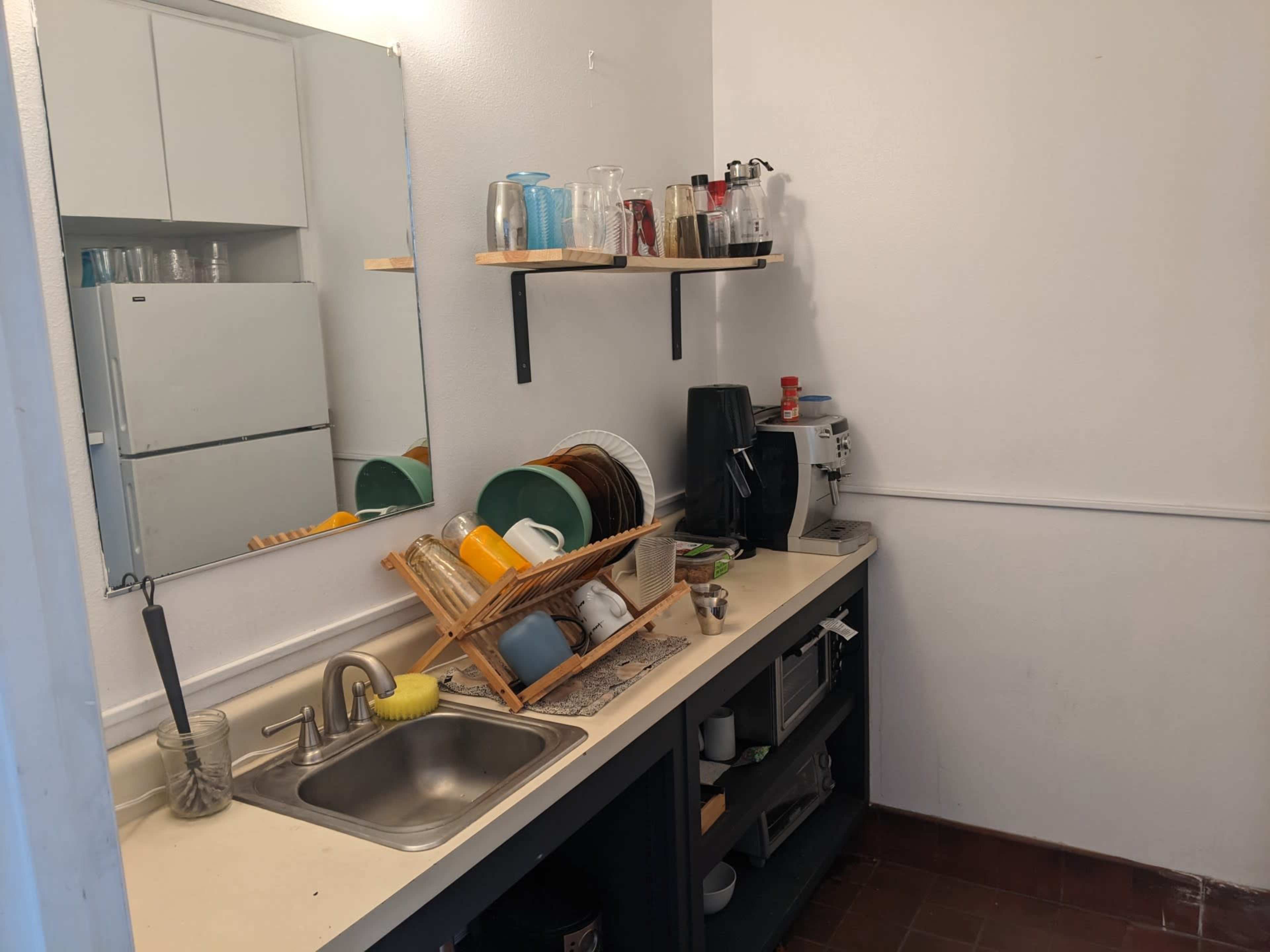 A small kitchen area with a sink, shelves holding glasses and dishes, and a refrigerator in the background.