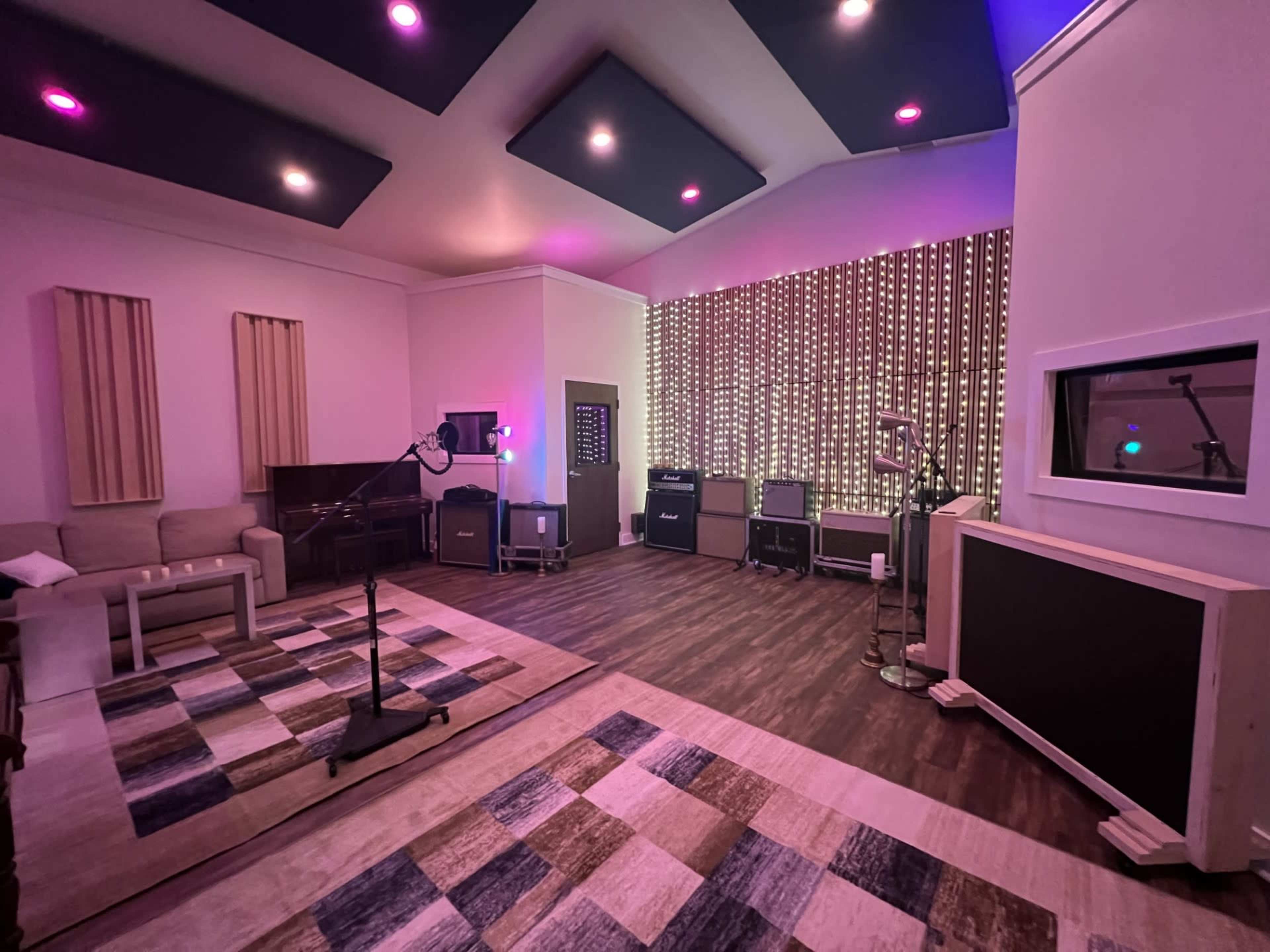 A music studio featuring a microphone stand, several amplifiers, and colorful ambient lighting on the walls and ceiling.