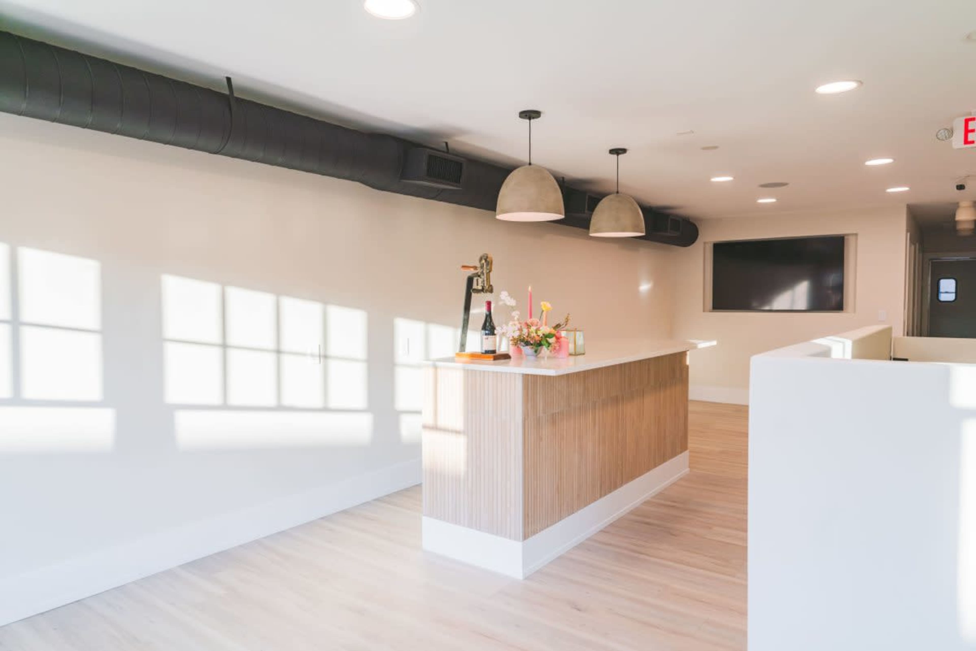A modern reception area with a light wood and white counter, decorative flowers, and overhead lighting, featuring large windows casting shadows on the walls.