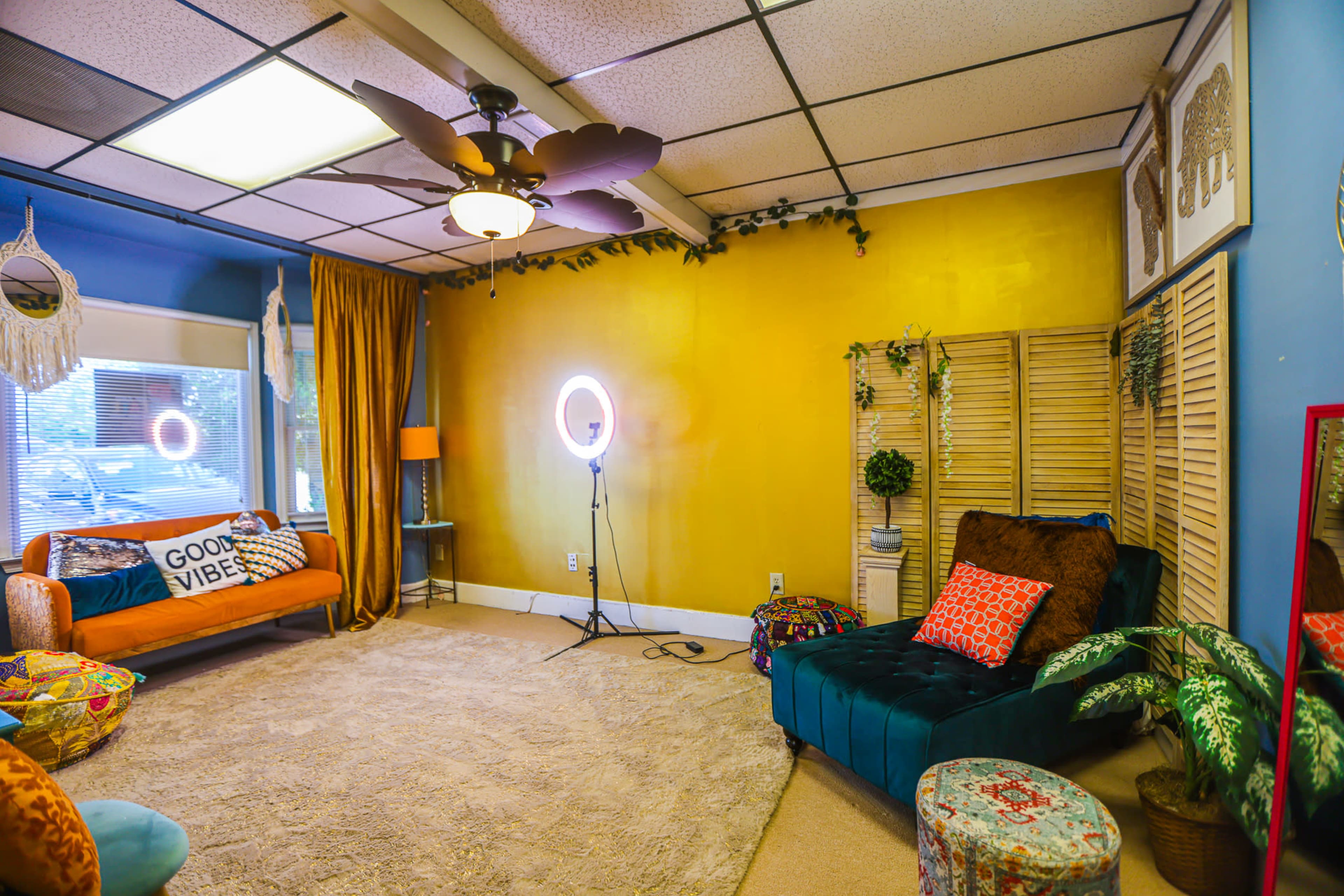 The image shows a cozy room with a yellow accent wall, a circular light setup, a green velvet chair, and a bright orange couch, all surrounded by plants and decorative items.