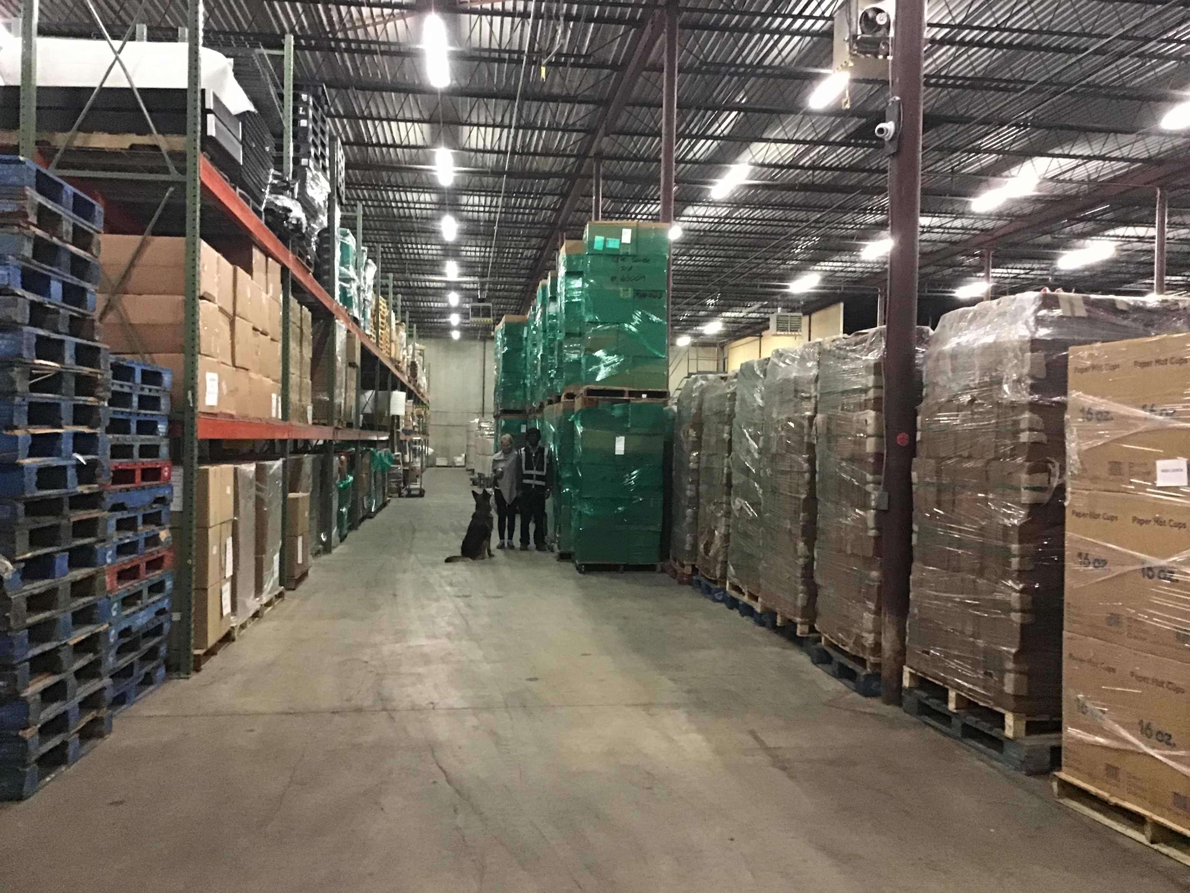 A warehouse interior shows stacked pallets of boxes and packaged goods arranged along both sides of the aisle.