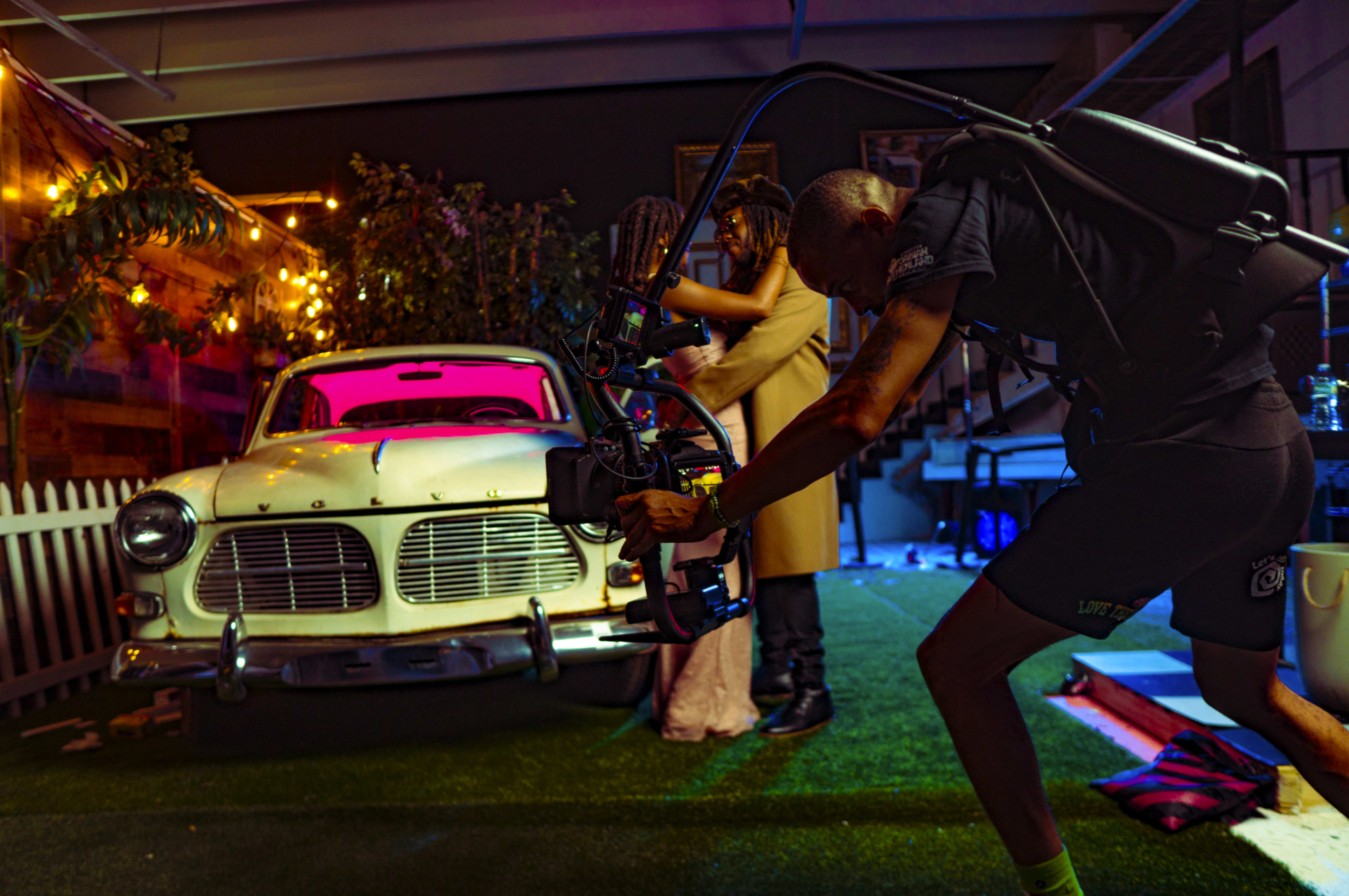 A man with a camera stabilization rig films a couple dancing near a vintage car in a dimly lit indoor setting decorated with lights.