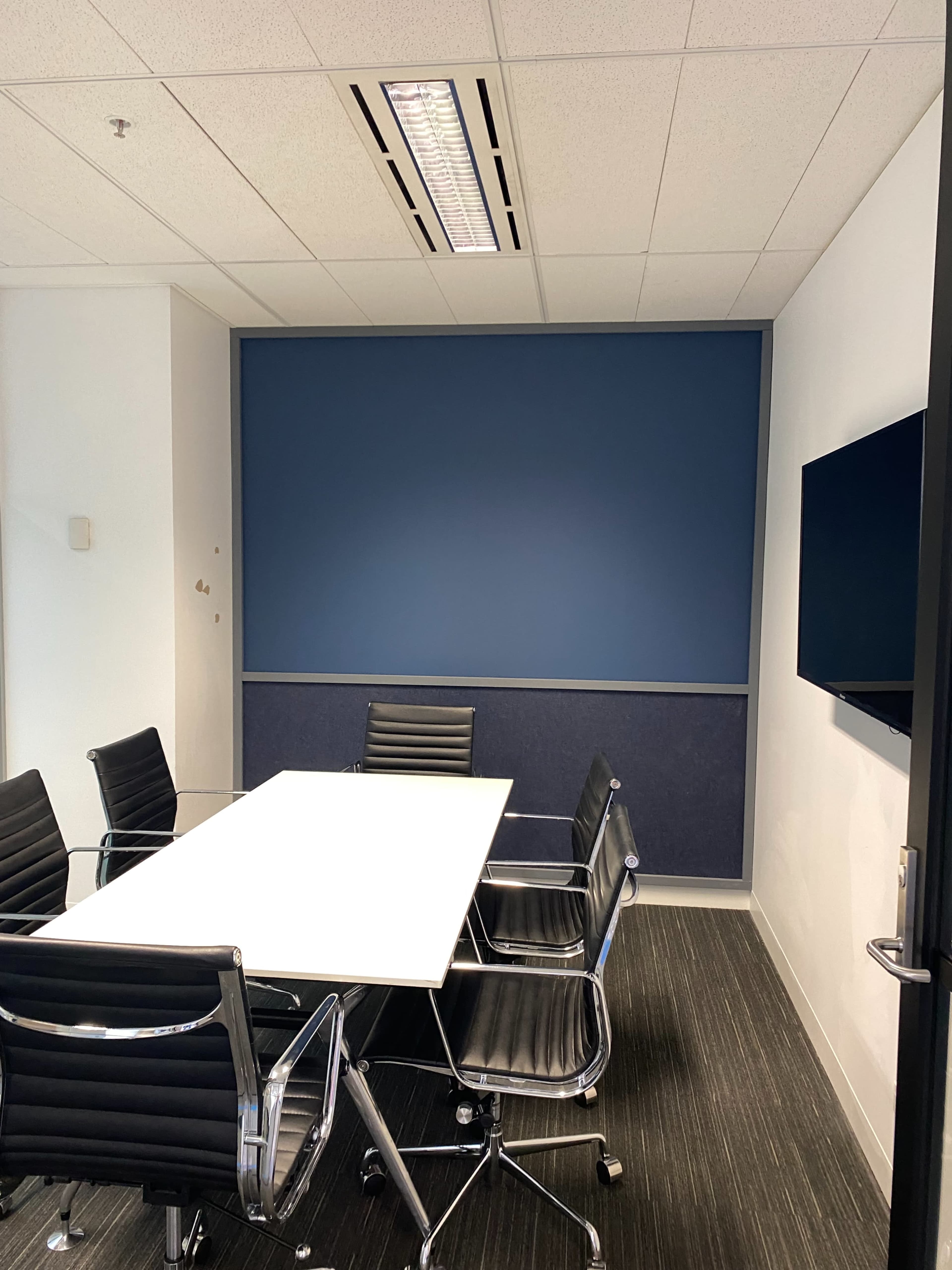 The image shows a conference room with a long white table surrounded by black chairs, a blue accent wall, and a wall-mounted television.