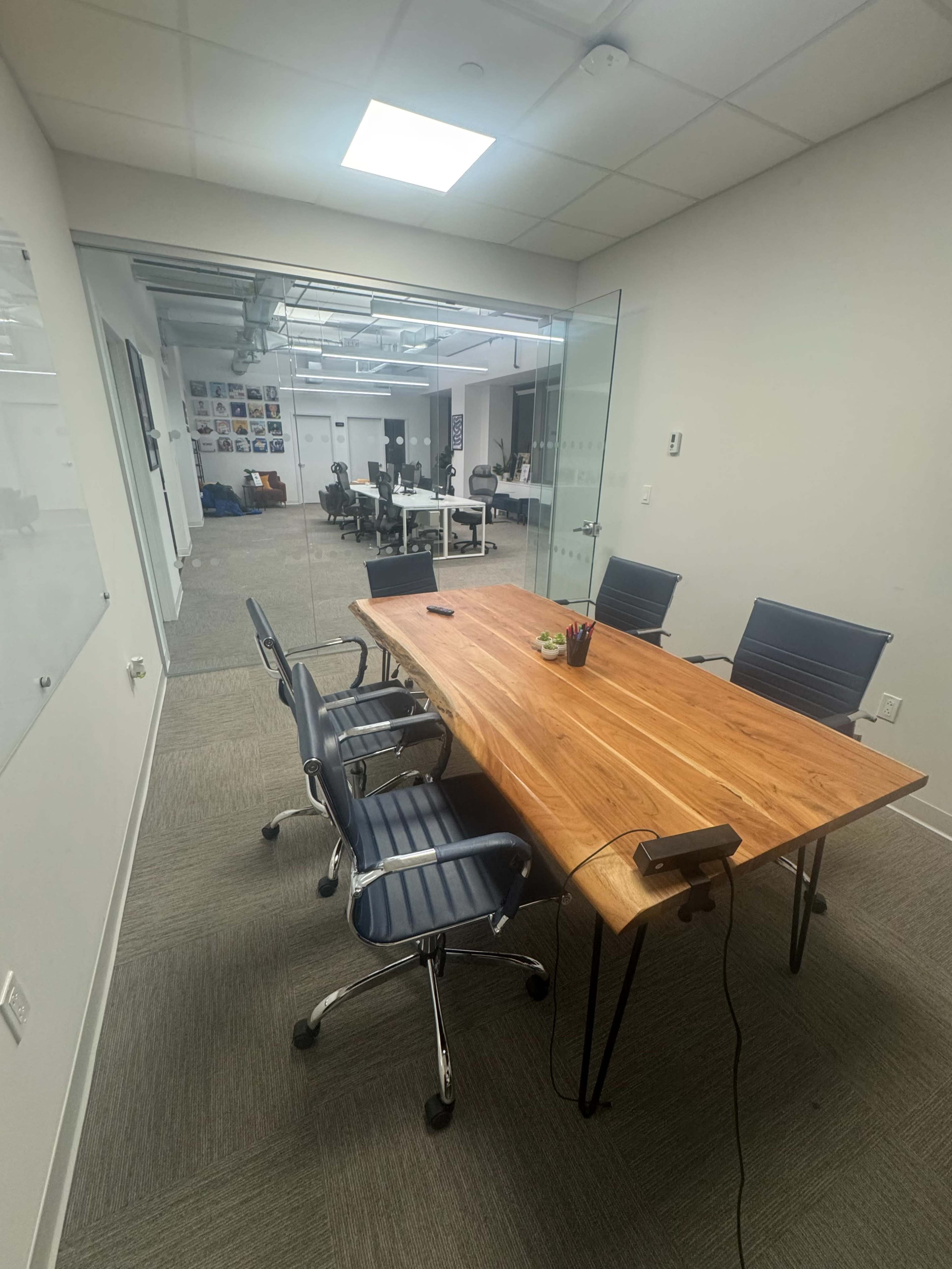 A conference room features a wooden table surrounded by four chairs, with glass walls revealing a workspace beyond.
