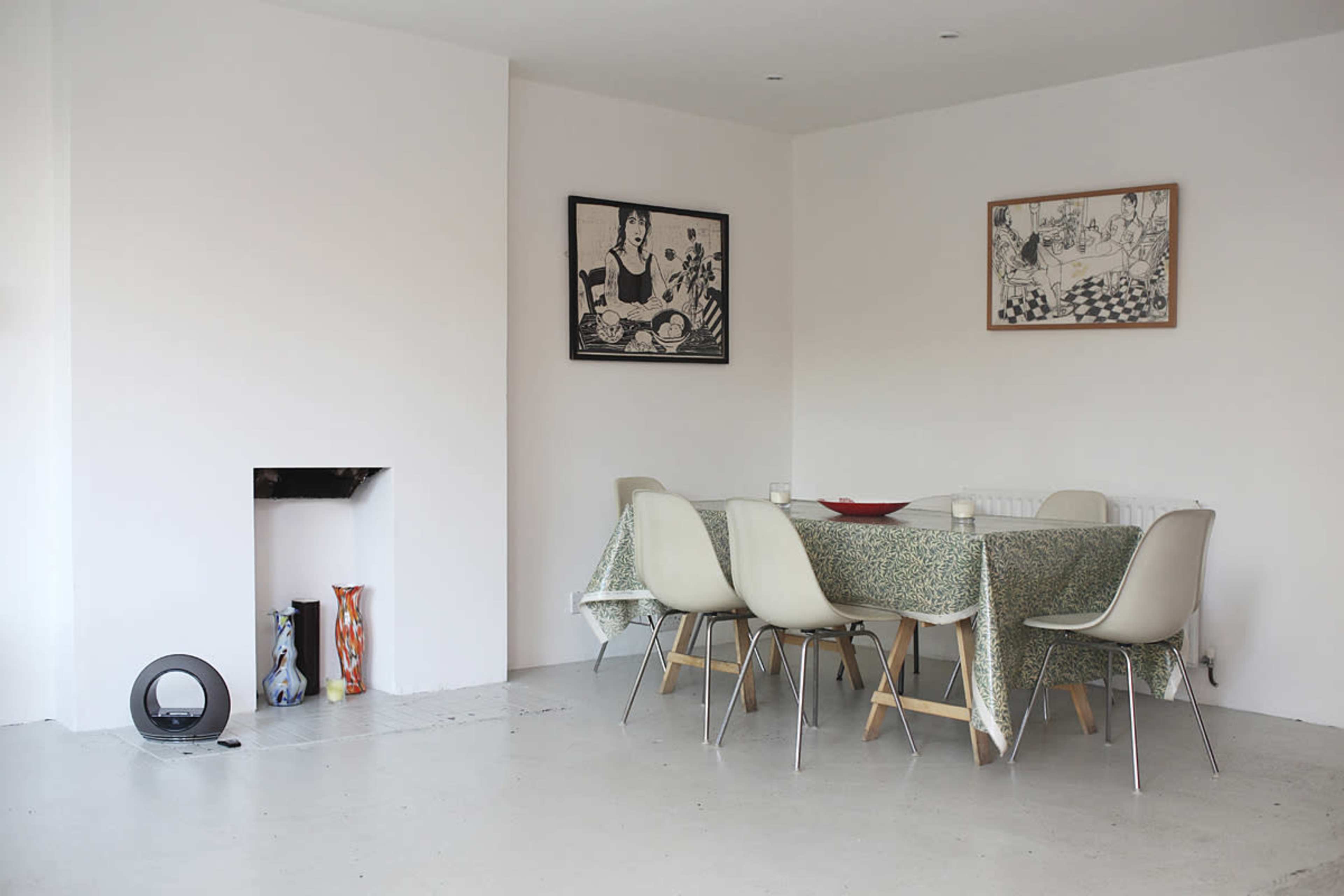A simple dining area features a wooden table with a patterned cloth, surrounded by several chairs, and adorned with art on the walls.