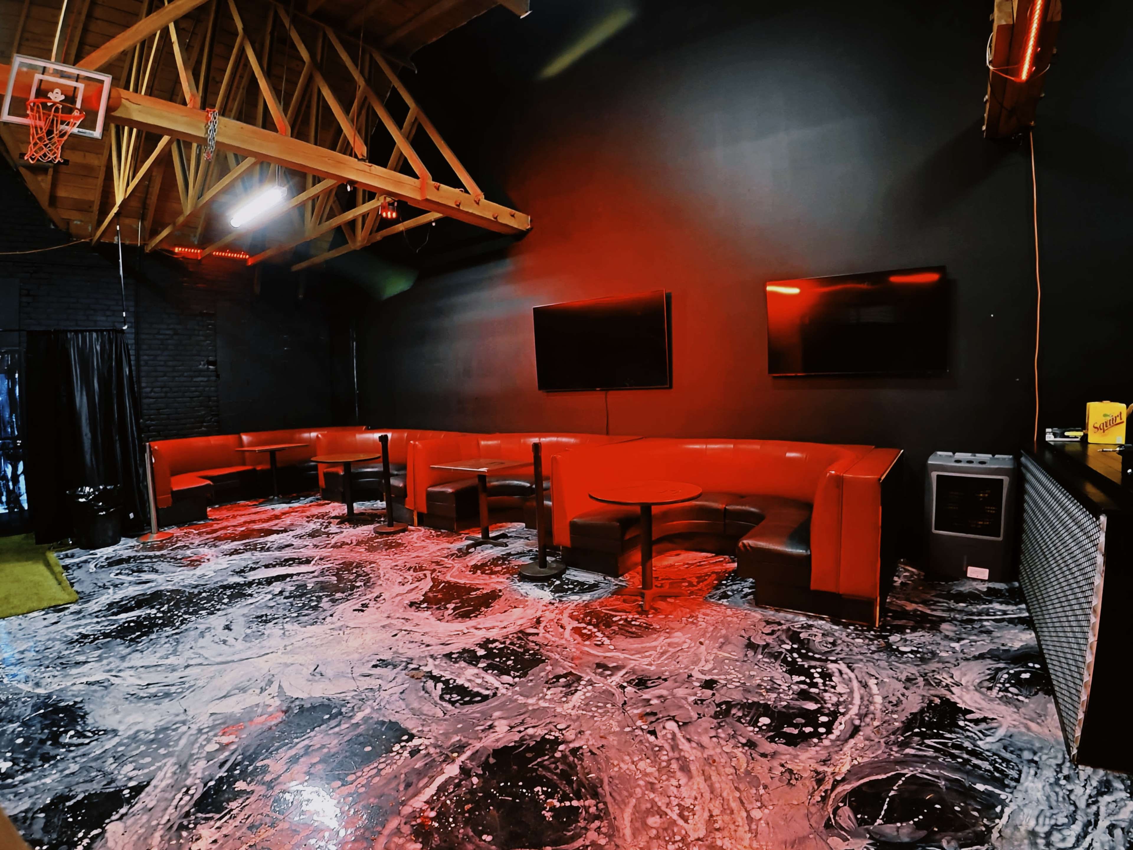 The image shows a dimly lit lounge area with a black wall, red seating, and two flat-screen TVs mounted on the wall.
