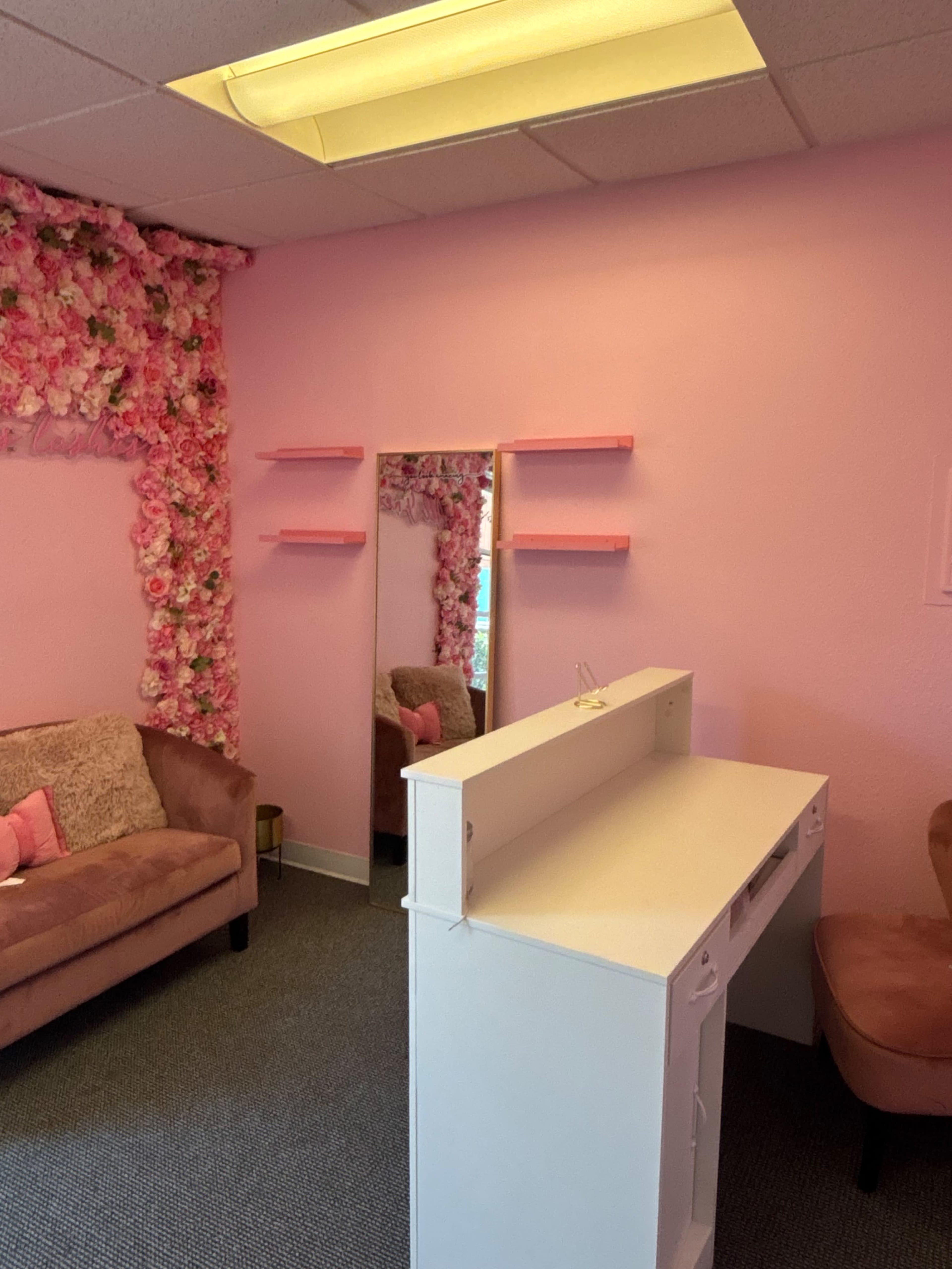 The image shows a pink-themed room decorated with floral accents, featuring a sofa, a mirror, and a white desk with shelves.