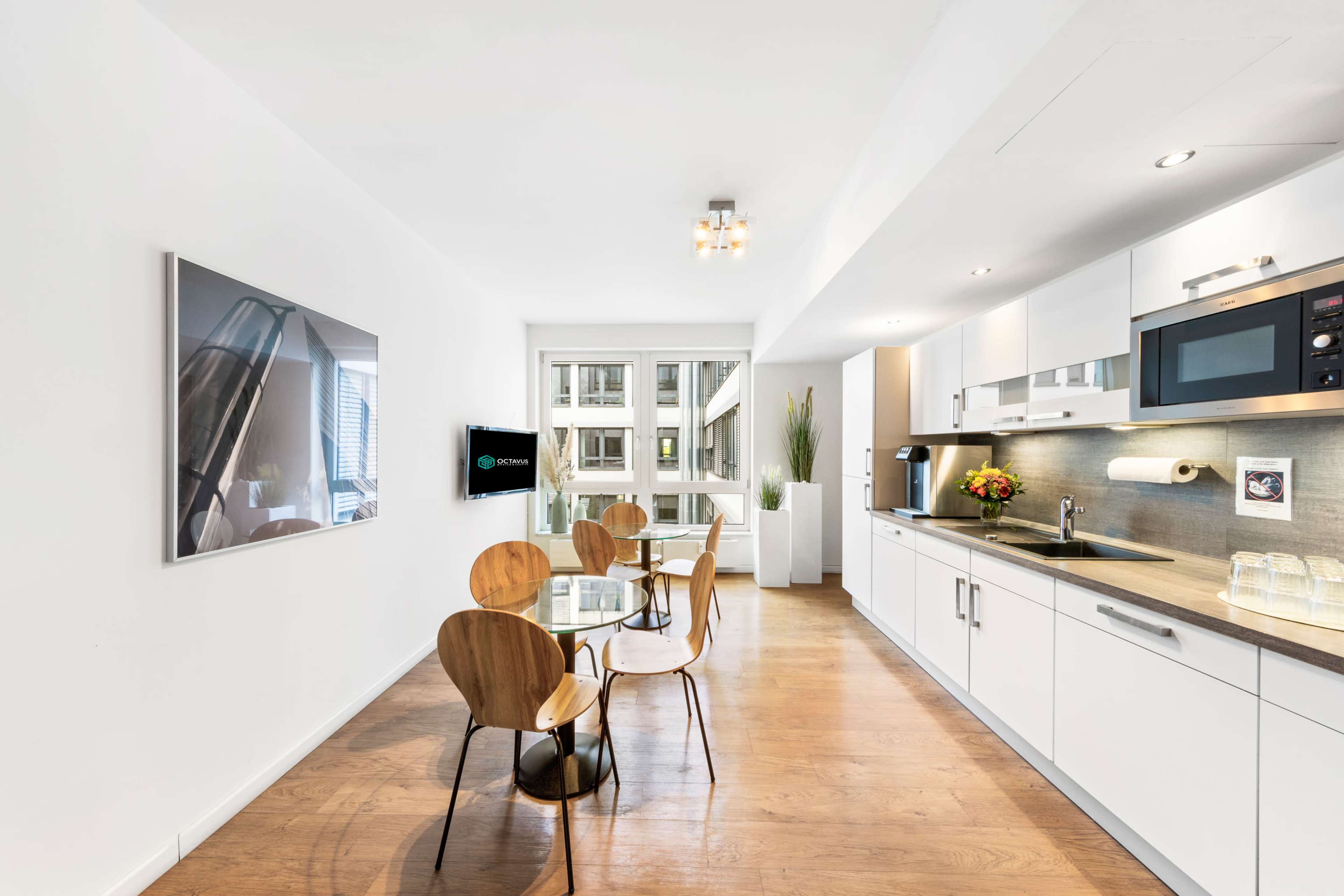 The image depicts a modern kitchen and dining area featuring a round table with four wooden chairs, white cabinets, and large windows allowing natural light.