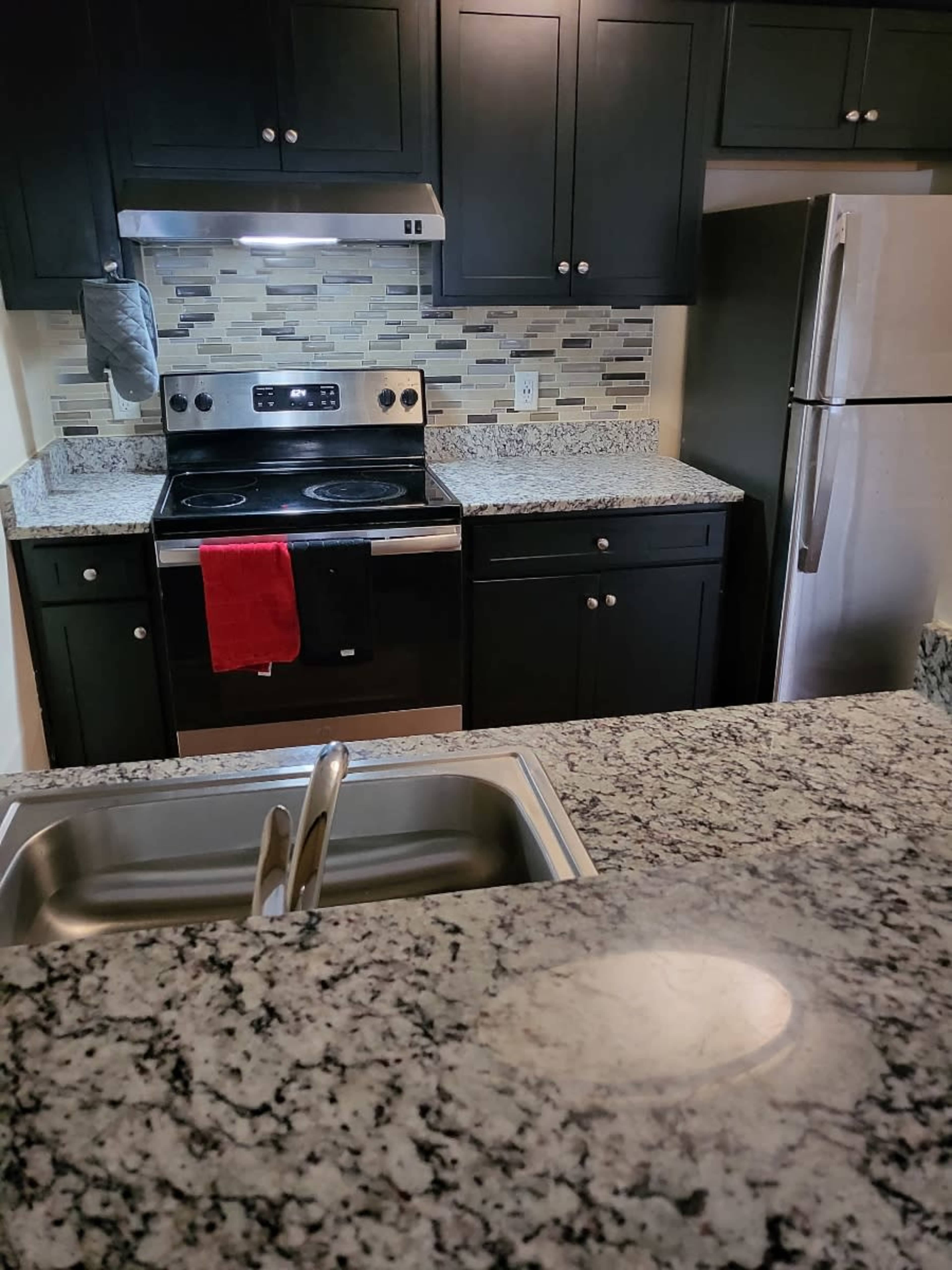 A kitchen with dark cabinets, granite countertops, a stainless steel refrigerator, and an oven with a red dish towel hanging from the handle.