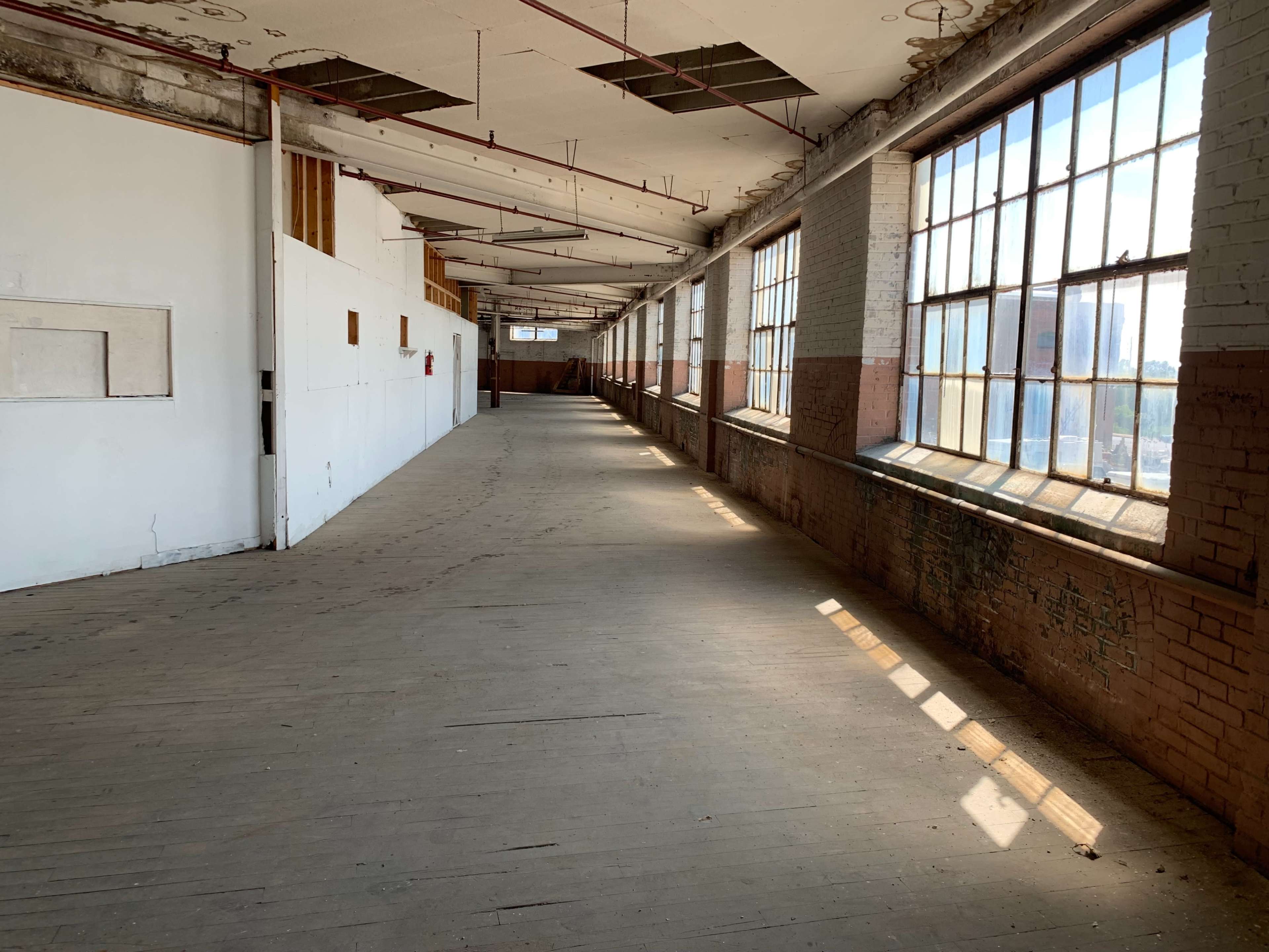 The image shows a long, empty corridor with large windows along one side, allowing sunlight to stream in onto the wooden floor.