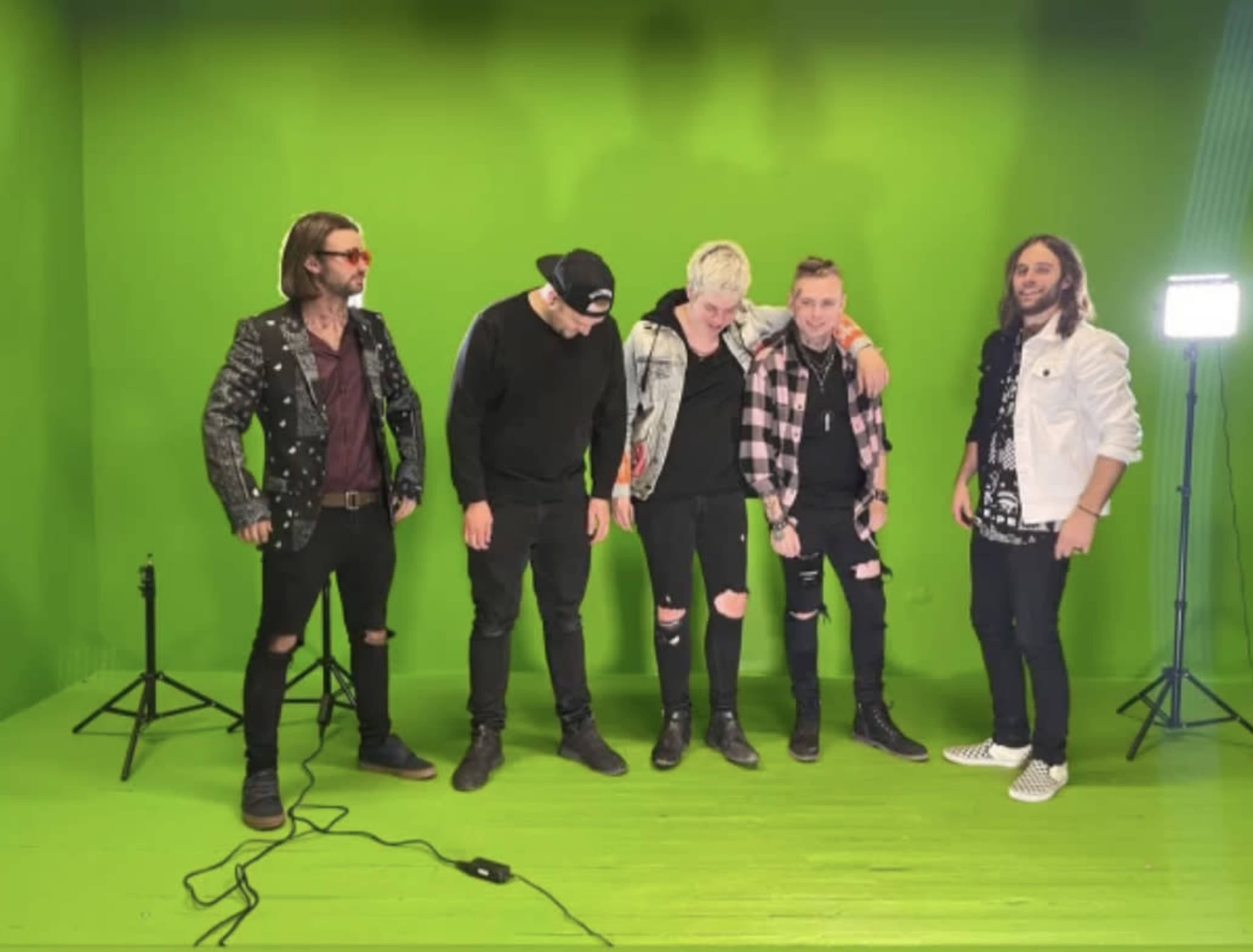 A group of five musicians stands in front of a green screen, dressed in casual, punk-style clothing.