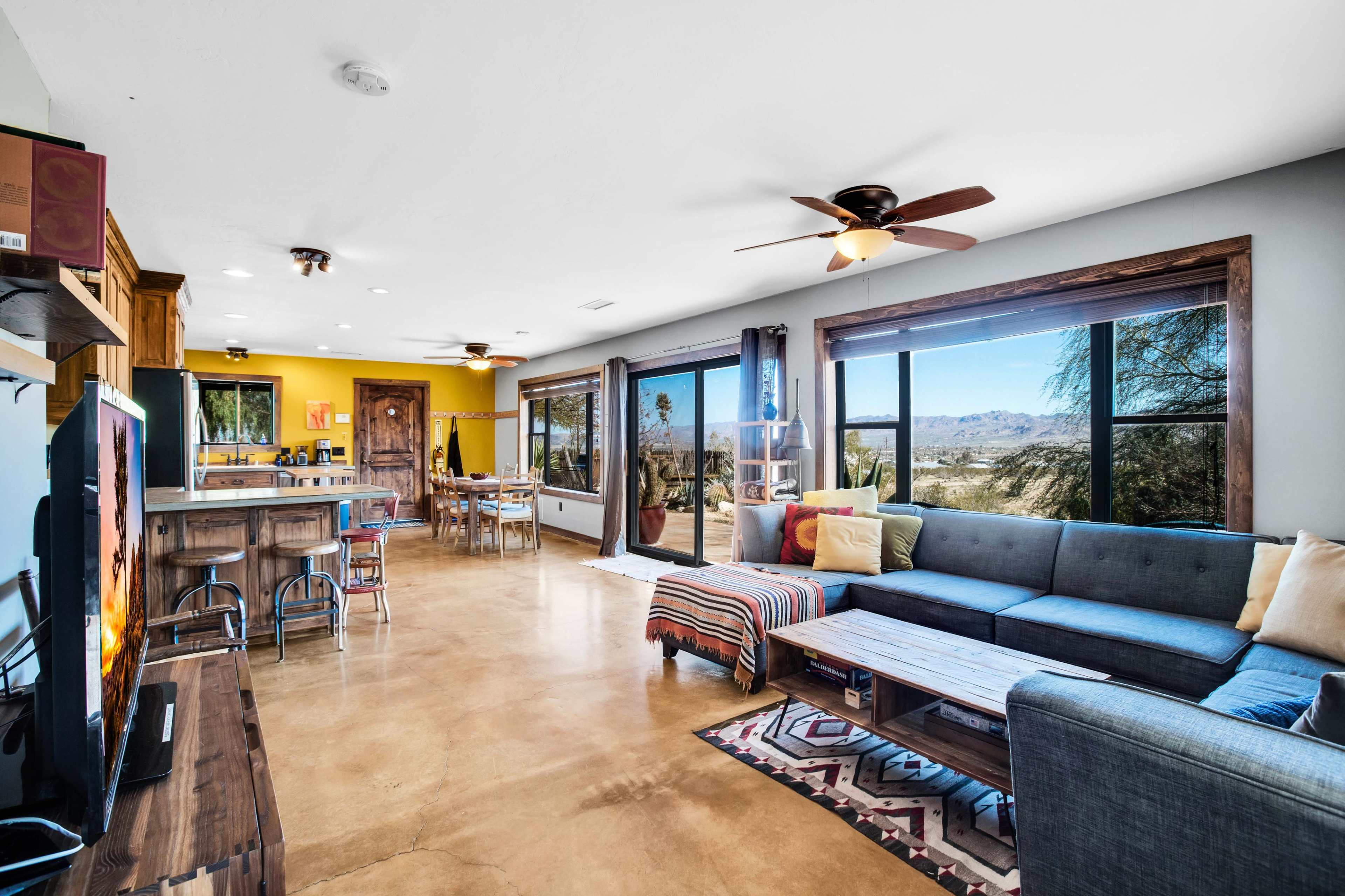 The living room features a modern interior with large windows offering a view of the outdoors, a sectional sofa, and a dining area.