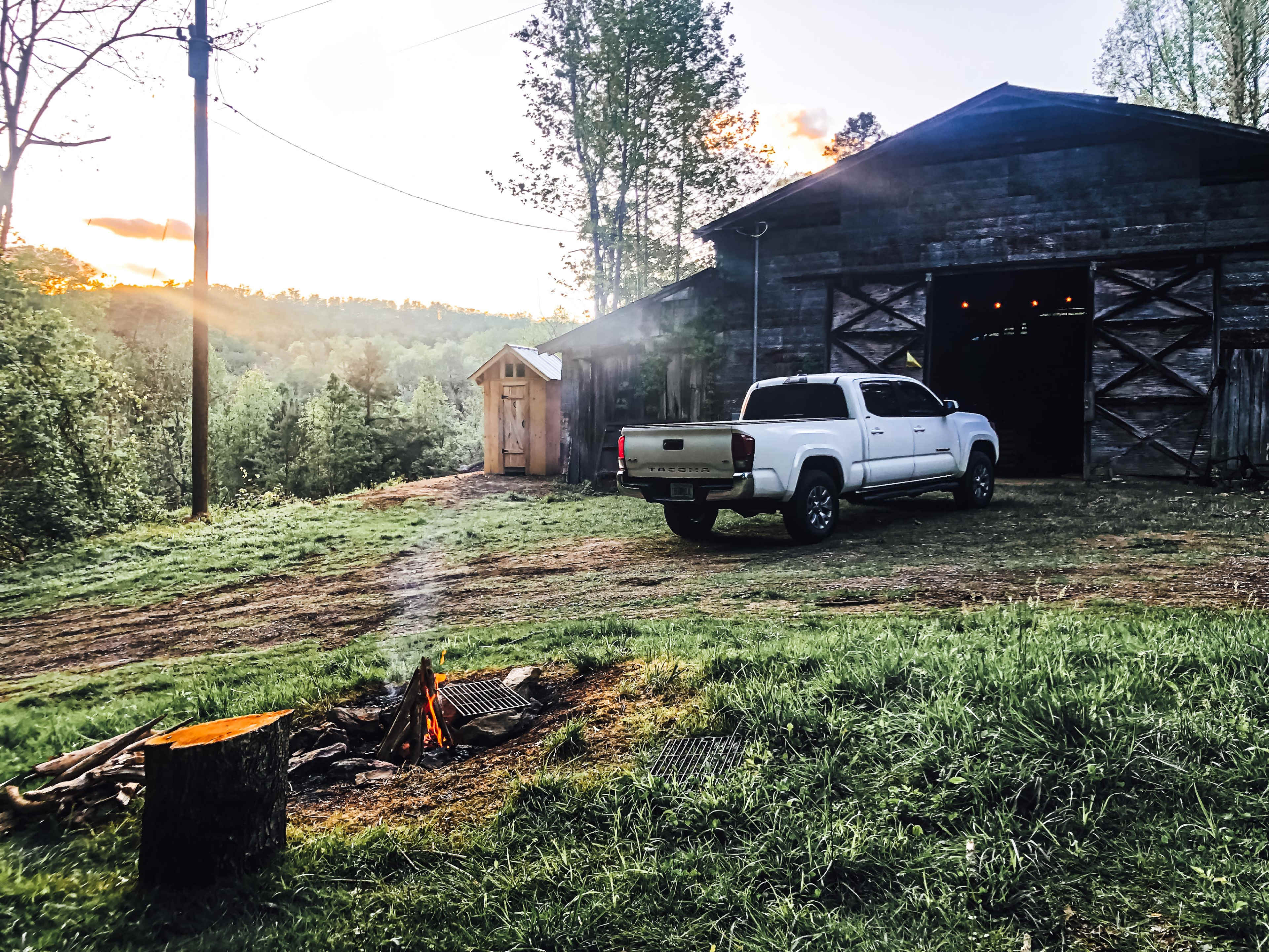 A white pickup truck is parked near a wooden barn at sunset, with a small outdoor fire pit emitting smoke in the foreground.