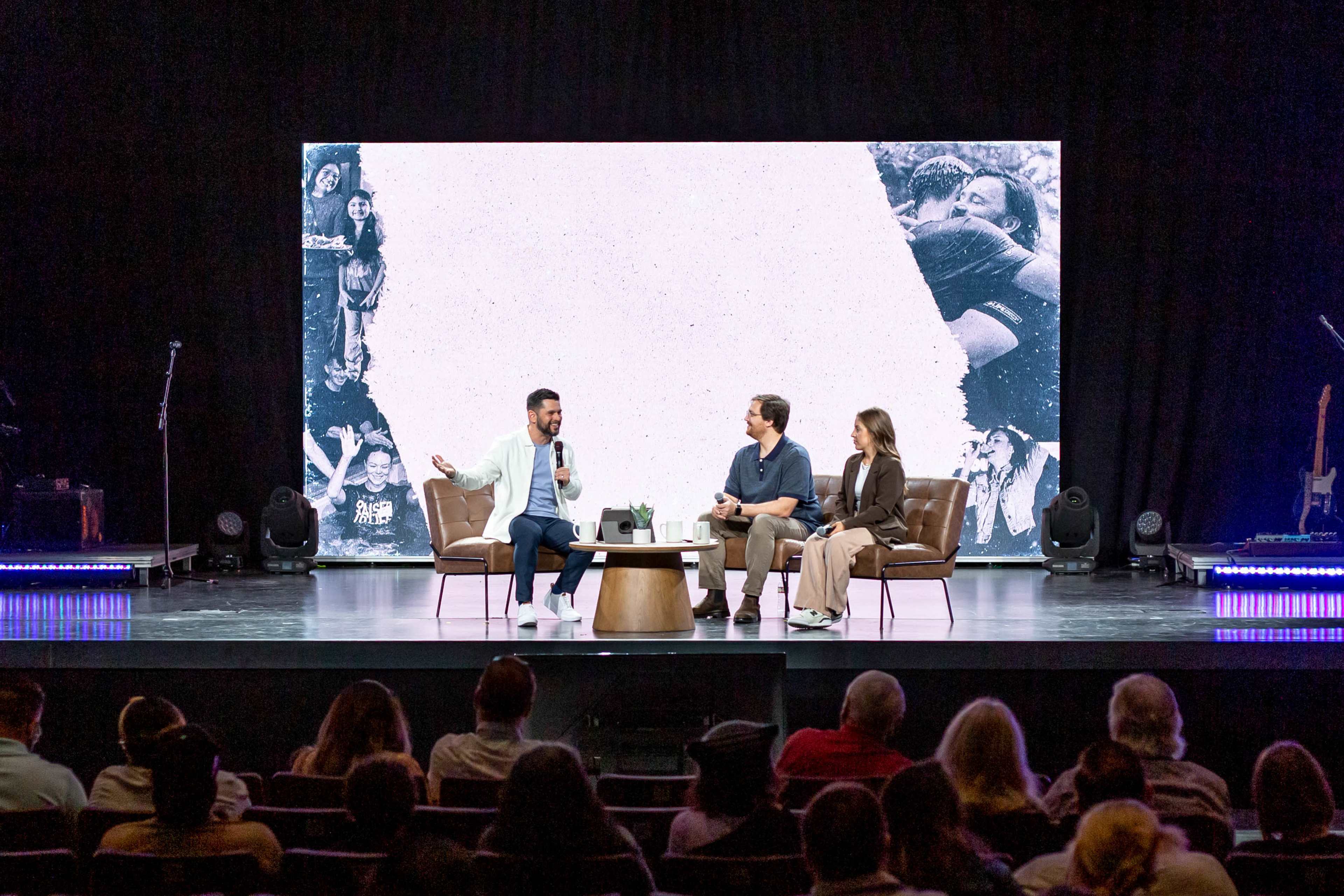 A panel discussion is taking place on a stage with two seated speakers and an audience in front, featuring a large backdrop displaying images of people.