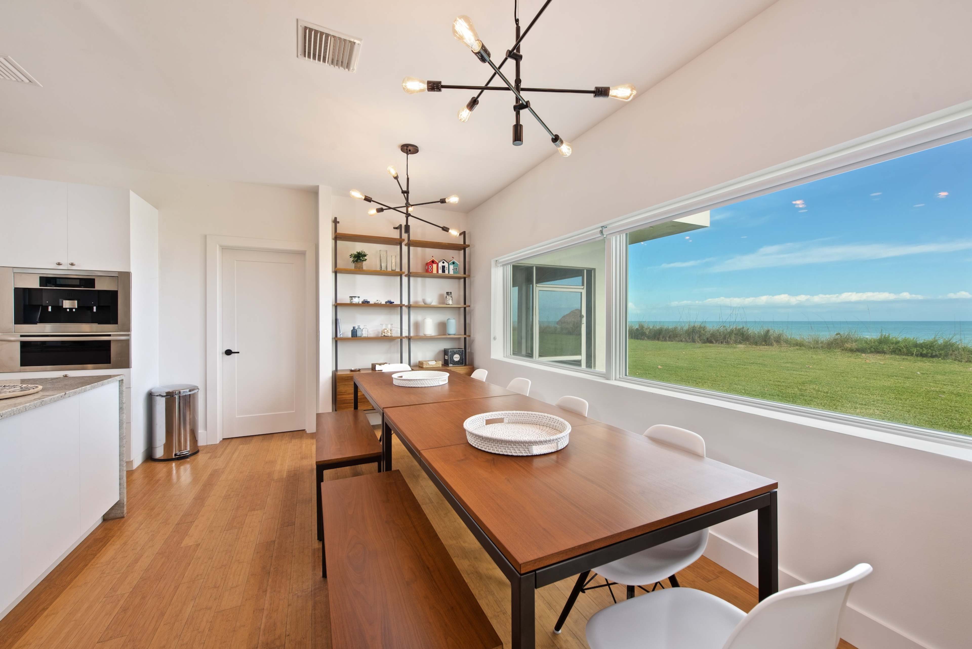 A modern dining room features a long wooden table surrounded by white chairs, with large windows offering a view of the ocean and green landscape outside.
