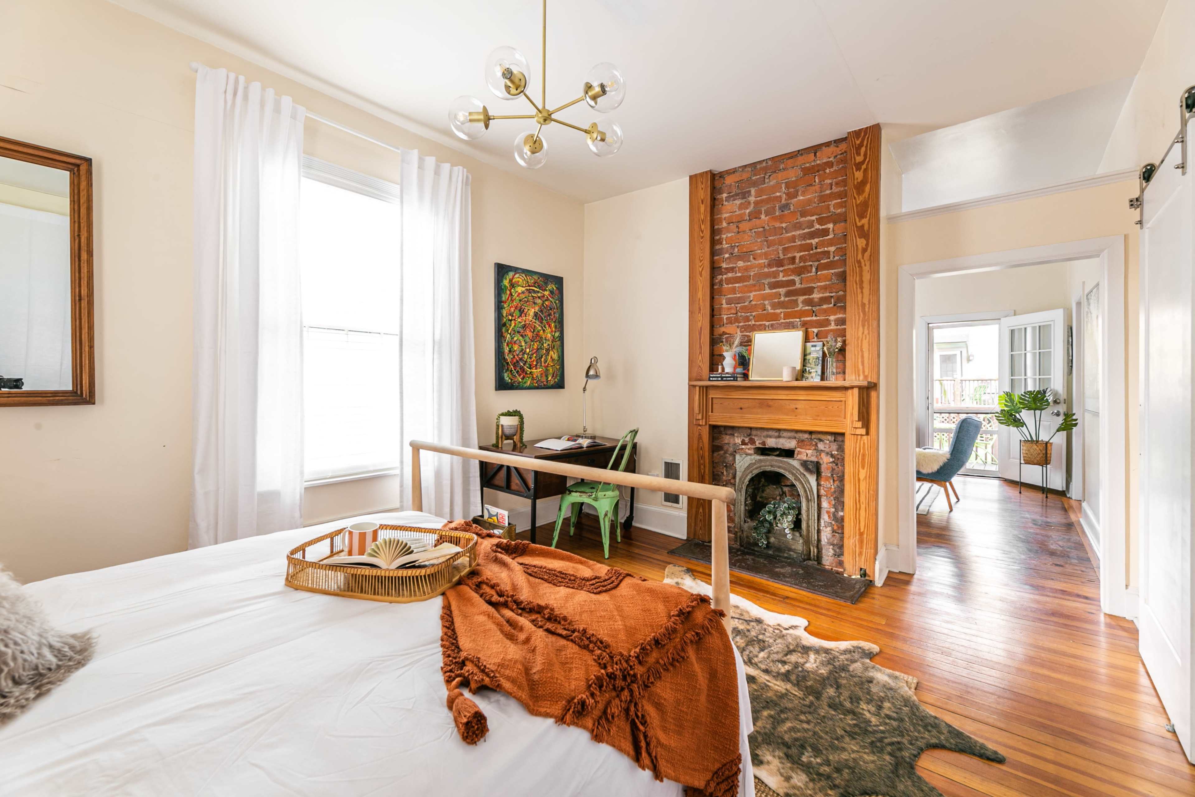 A cozy bedroom with a wooden bed, a brick fireplace, a small desk, and large windows with sheer white curtains.
