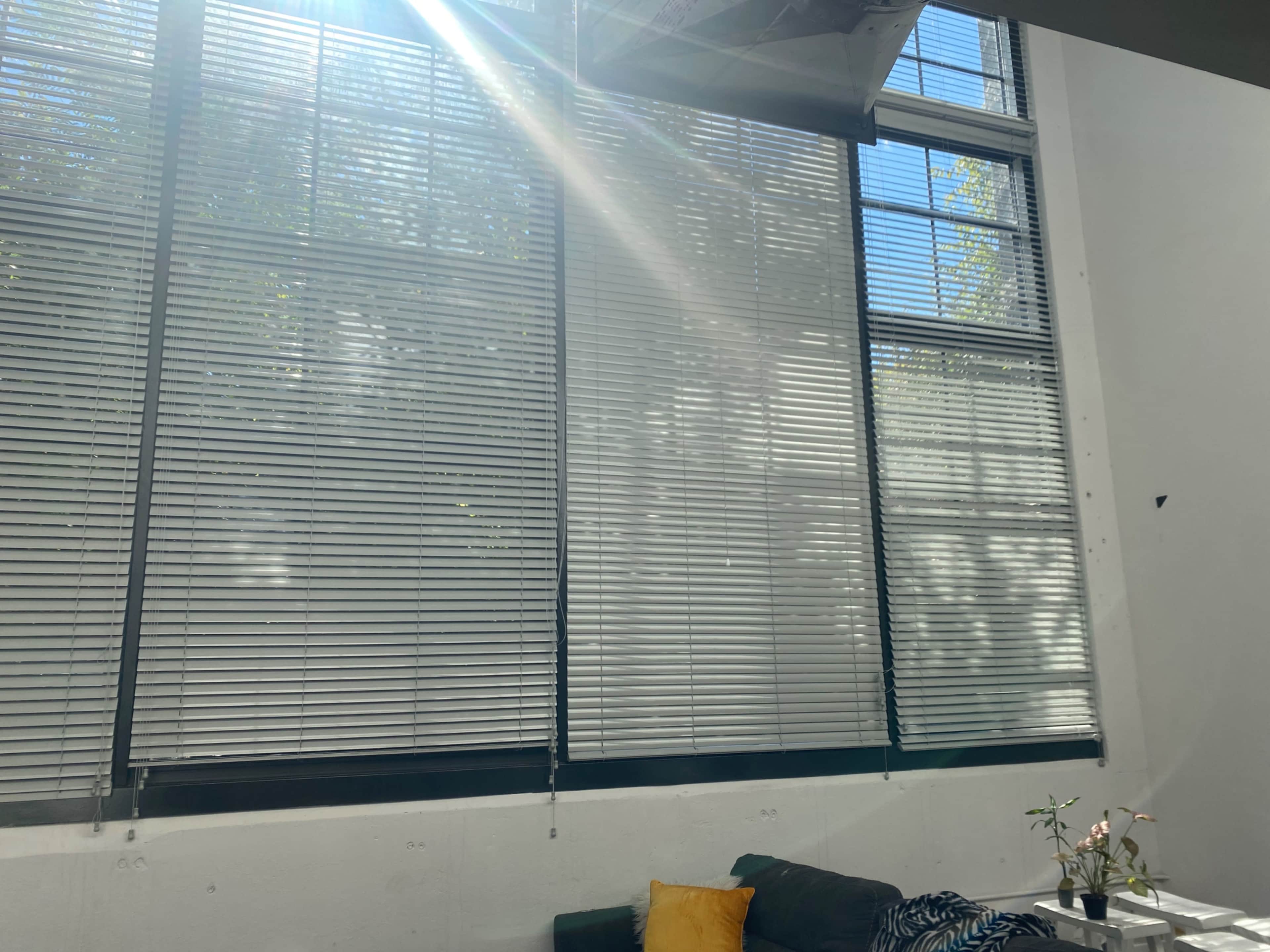 Large windows with blinds partially closed, allowing sunlight to stream into a room with a couch and a small table.