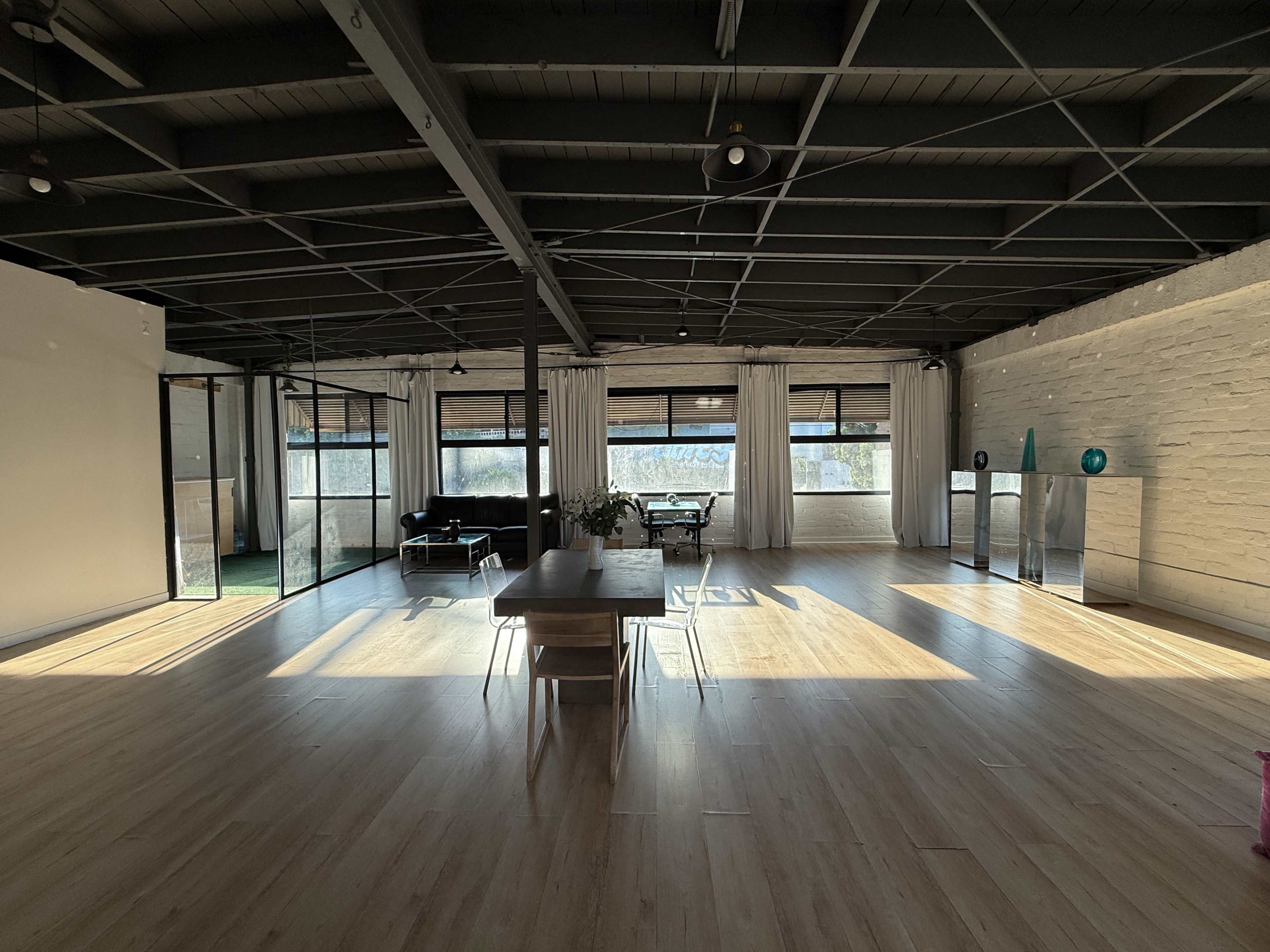 The image shows a spacious, modern interior with large windows, wooden flooring, and minimal furniture, including a table and chairs.