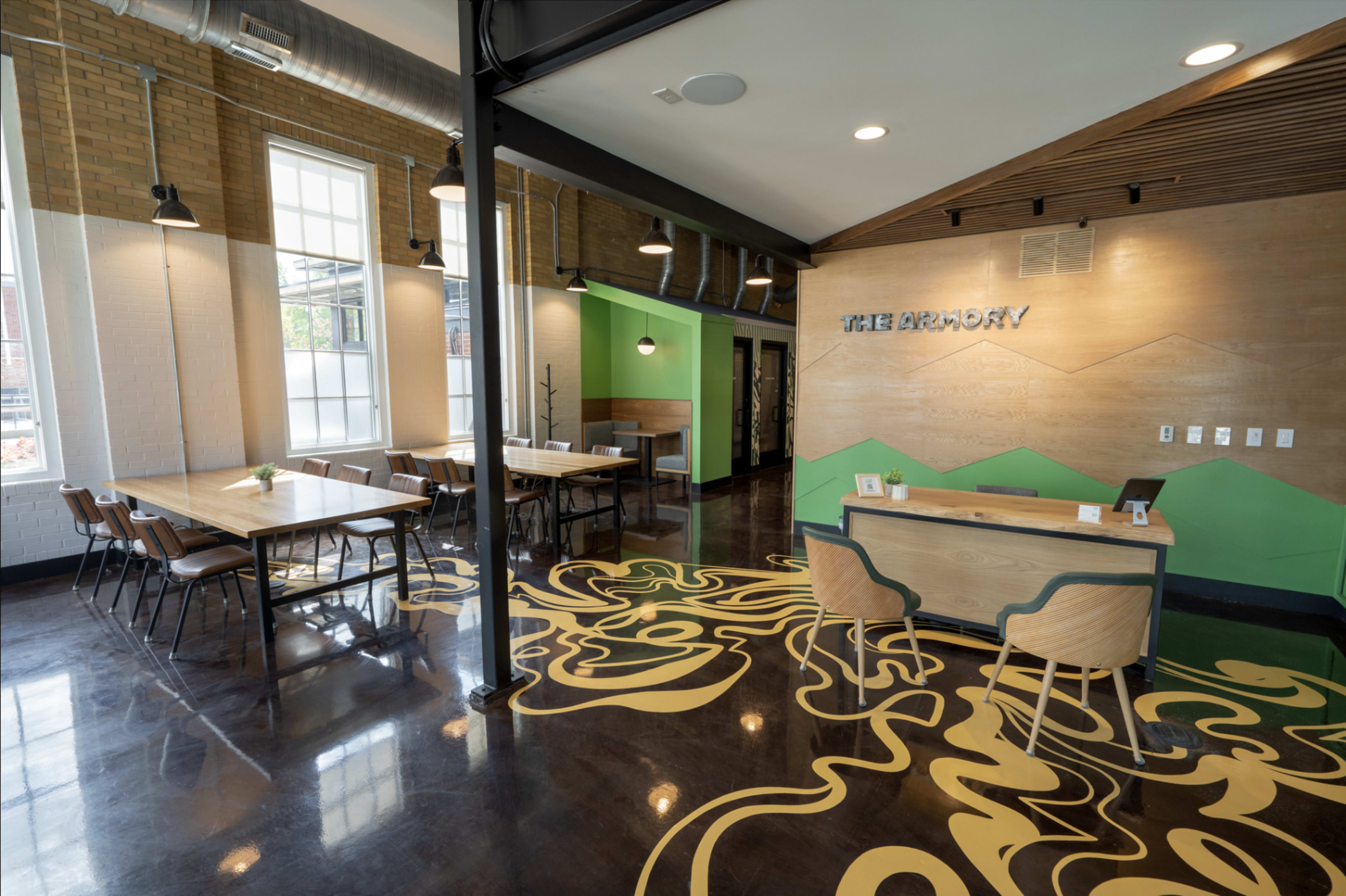 The image shows a modern interior space with wooden tables, chairs, and a reception desk, featuring green accent walls and a decorative floor design.