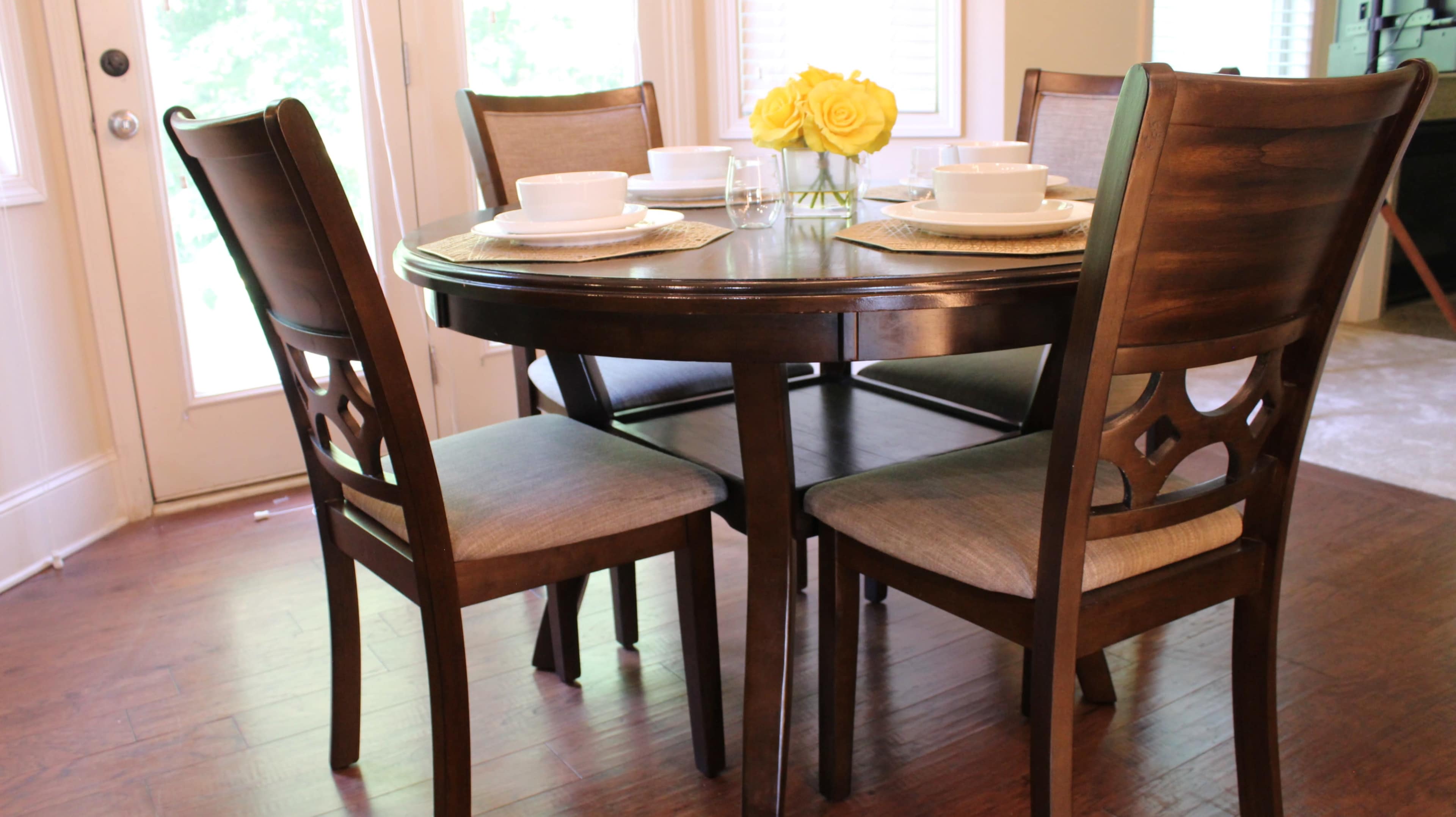 A round wooden dining table is set with white dishes and a vase of yellow roses, surrounded by four wooden chairs.