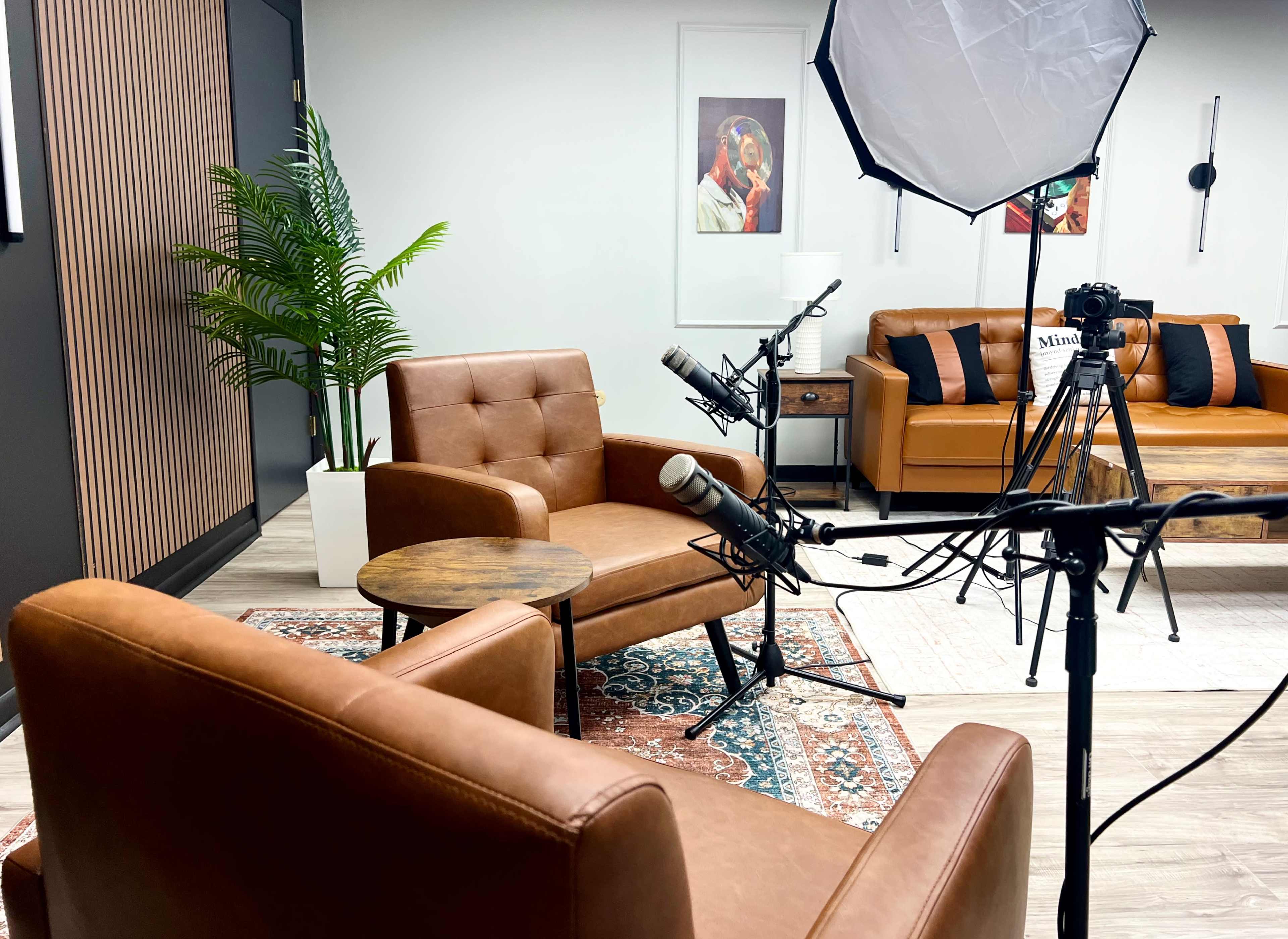 The image shows a modern, well-lit studio setup featuring two brown leather armchairs, a small round table, microphones, and a camera on a tripod, surrounded by decorative plants and artwork.