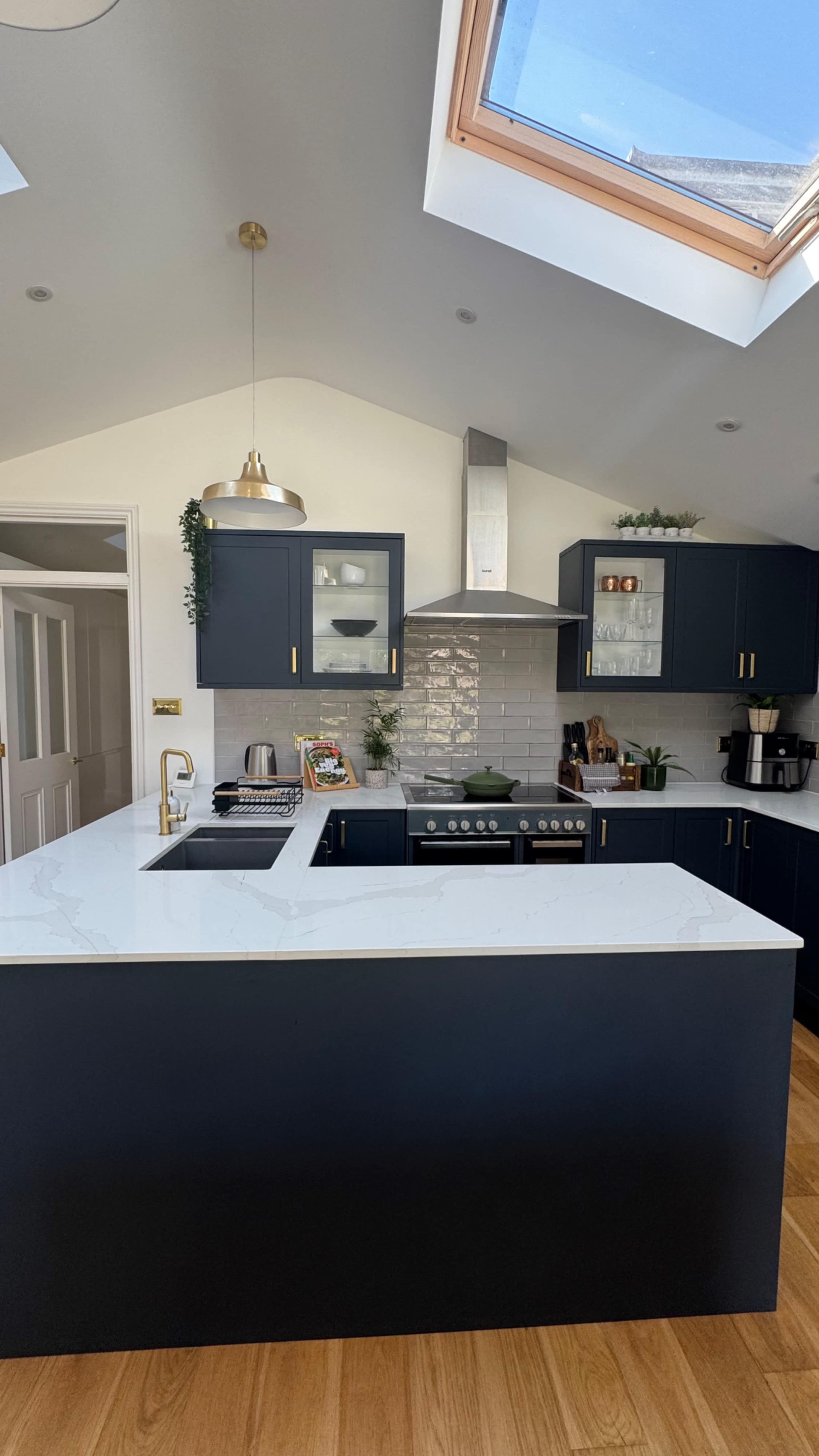 The image shows a modern kitchen with navy blue cabinets, a large white countertop, and a skylight allowing natural light to enter.