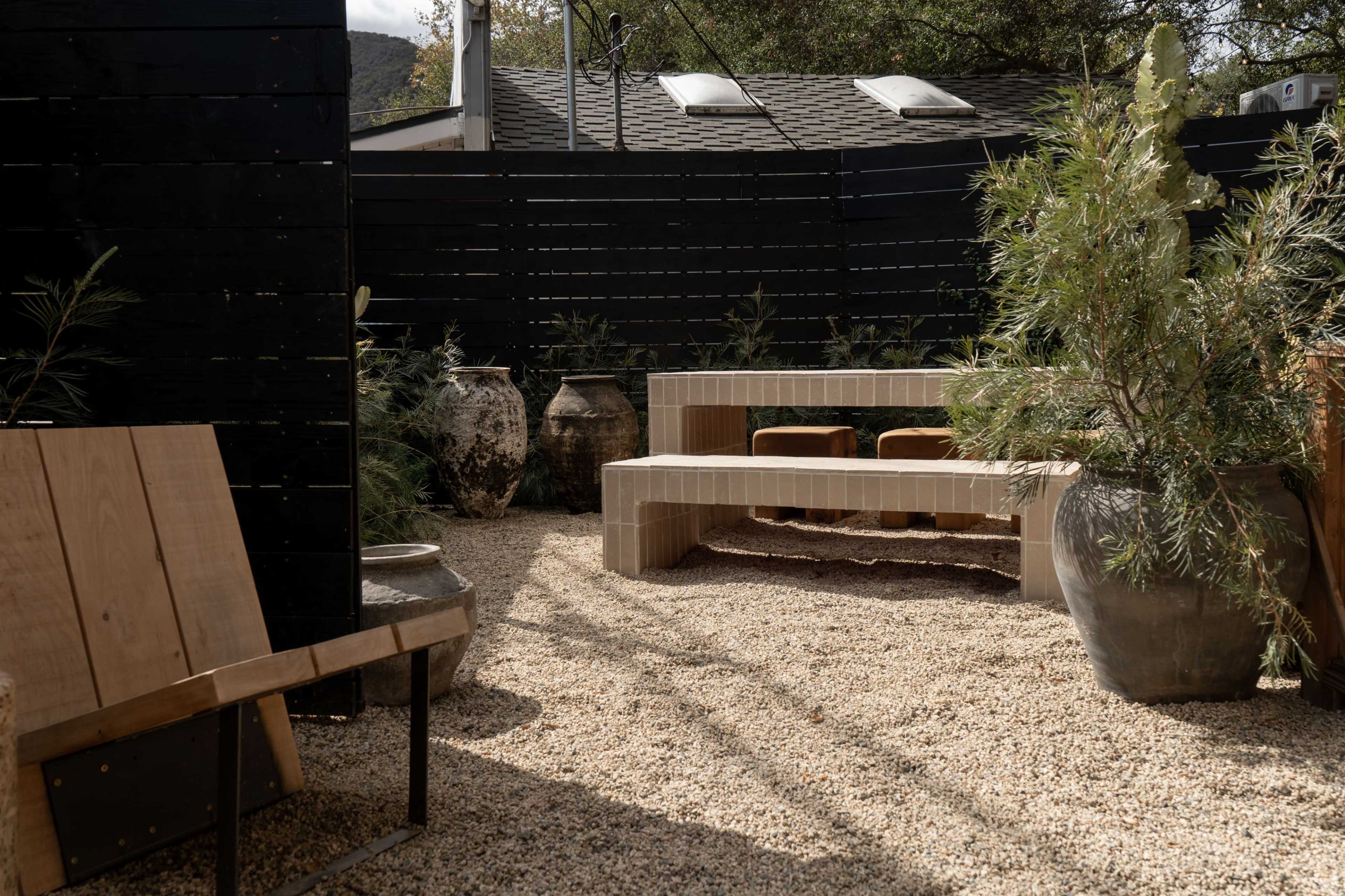 The image shows a gravel patio area featuring a minimalist bench, potted plants, and decorative pottery surrounded by a black wooden fence.