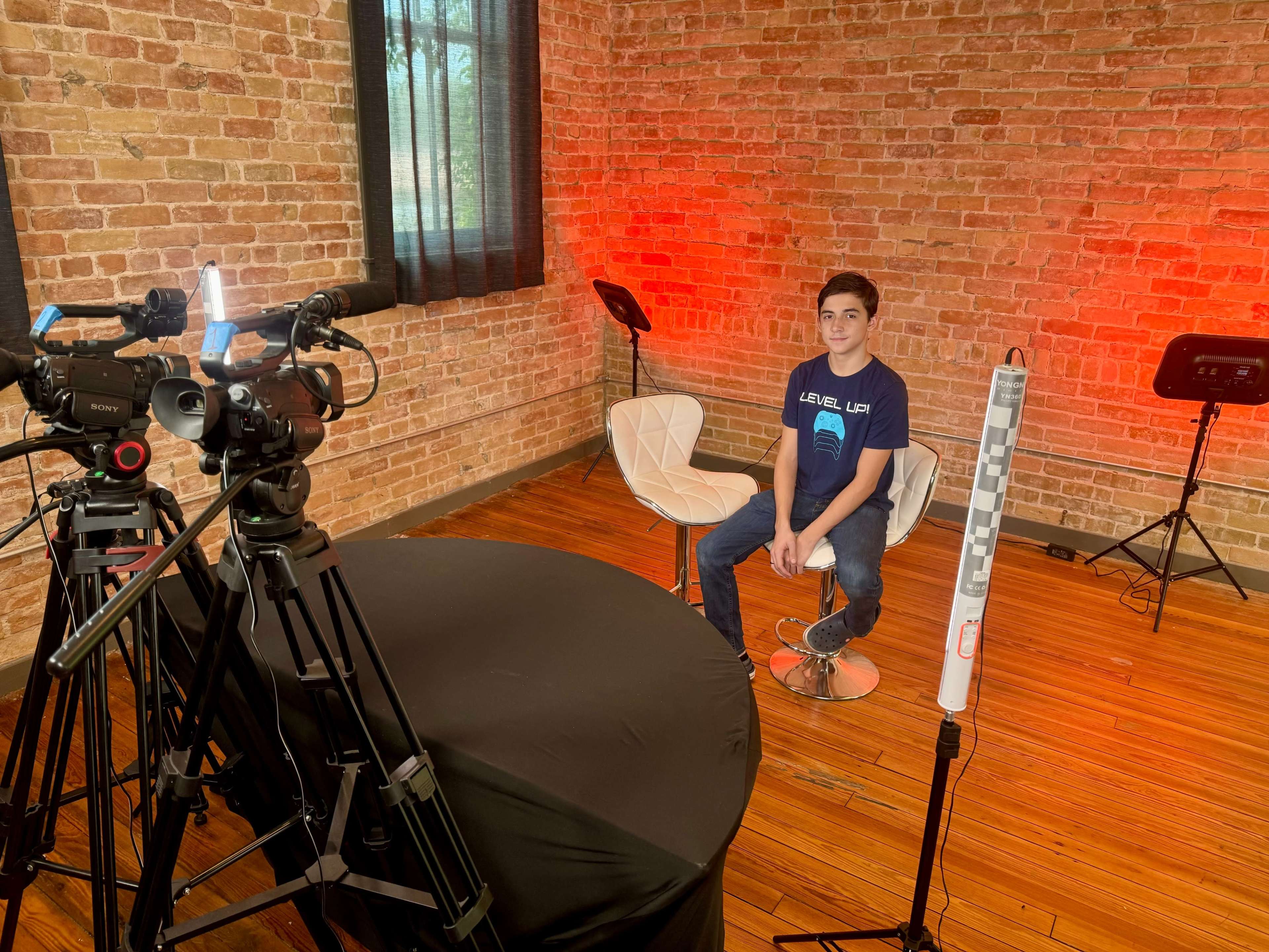 A young boy sits on a white chair in a brick-walled room set up for video recording, with cameras and equipment positioned around him.