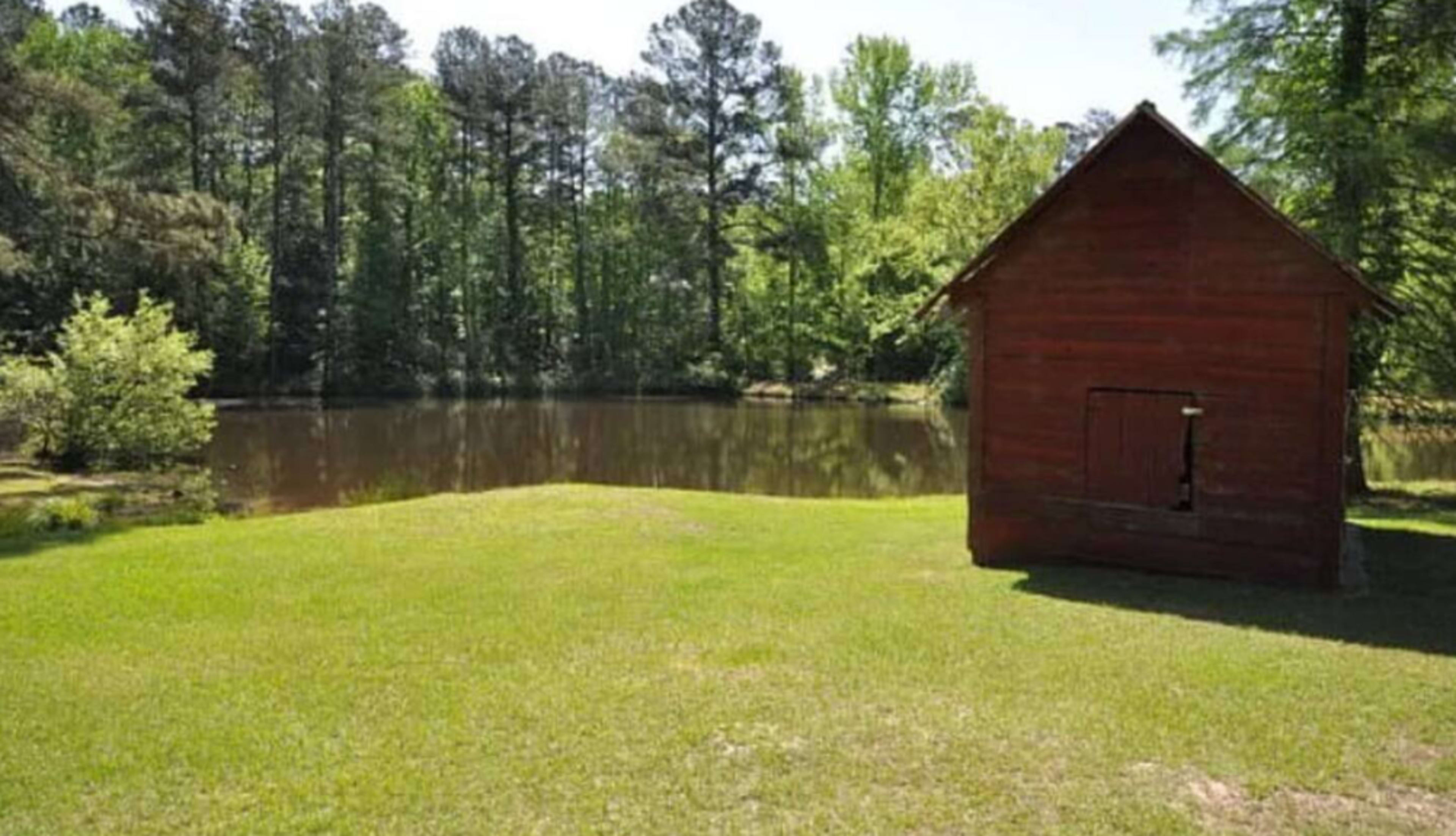 A small red wooden shed stands beside a calm pond surrounded by lush greenery.