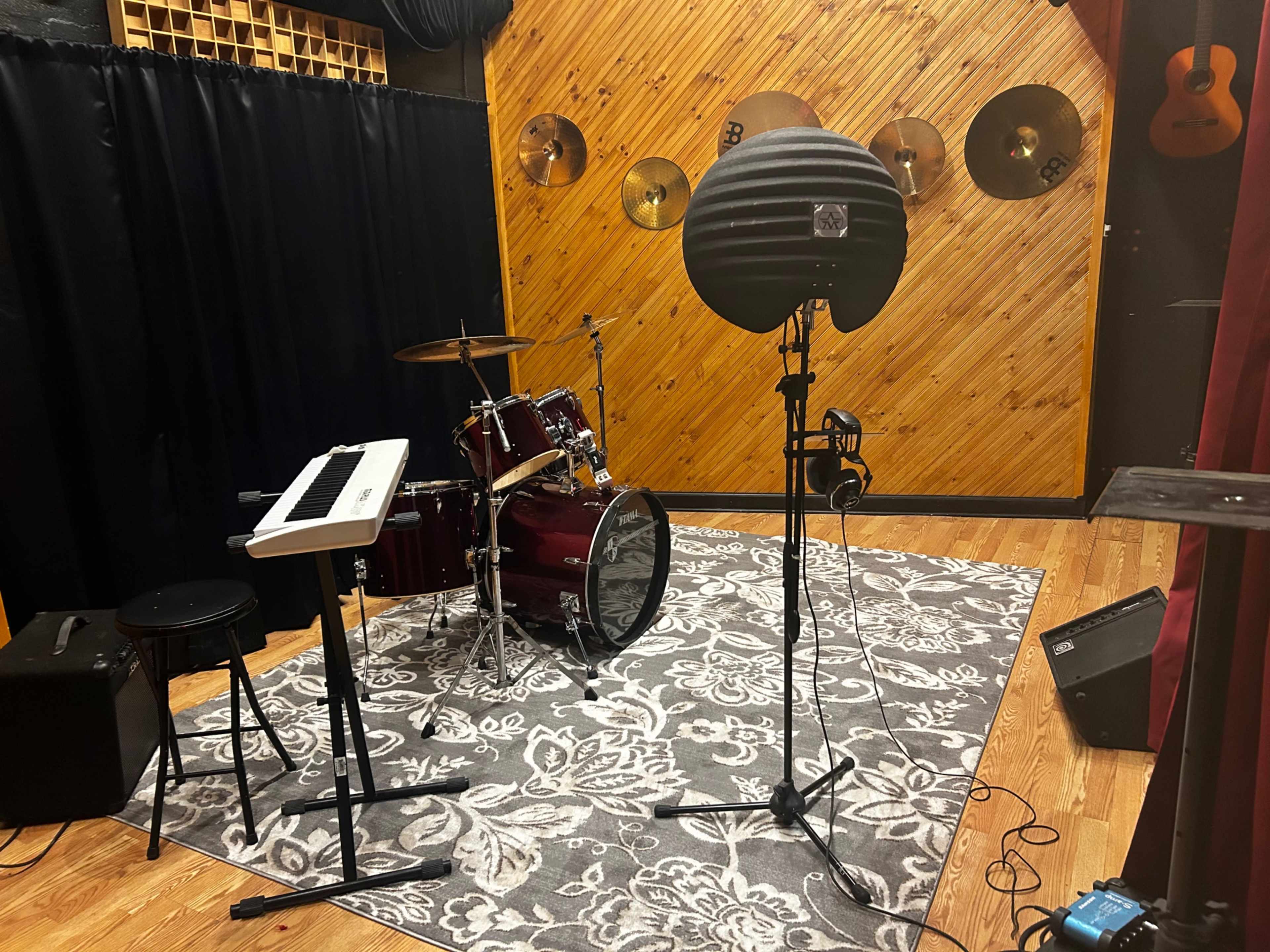 A music recording studio with a drum set, a keyboard on a stand, and a microphone setup surrounded by musical instruments and acoustic panels.